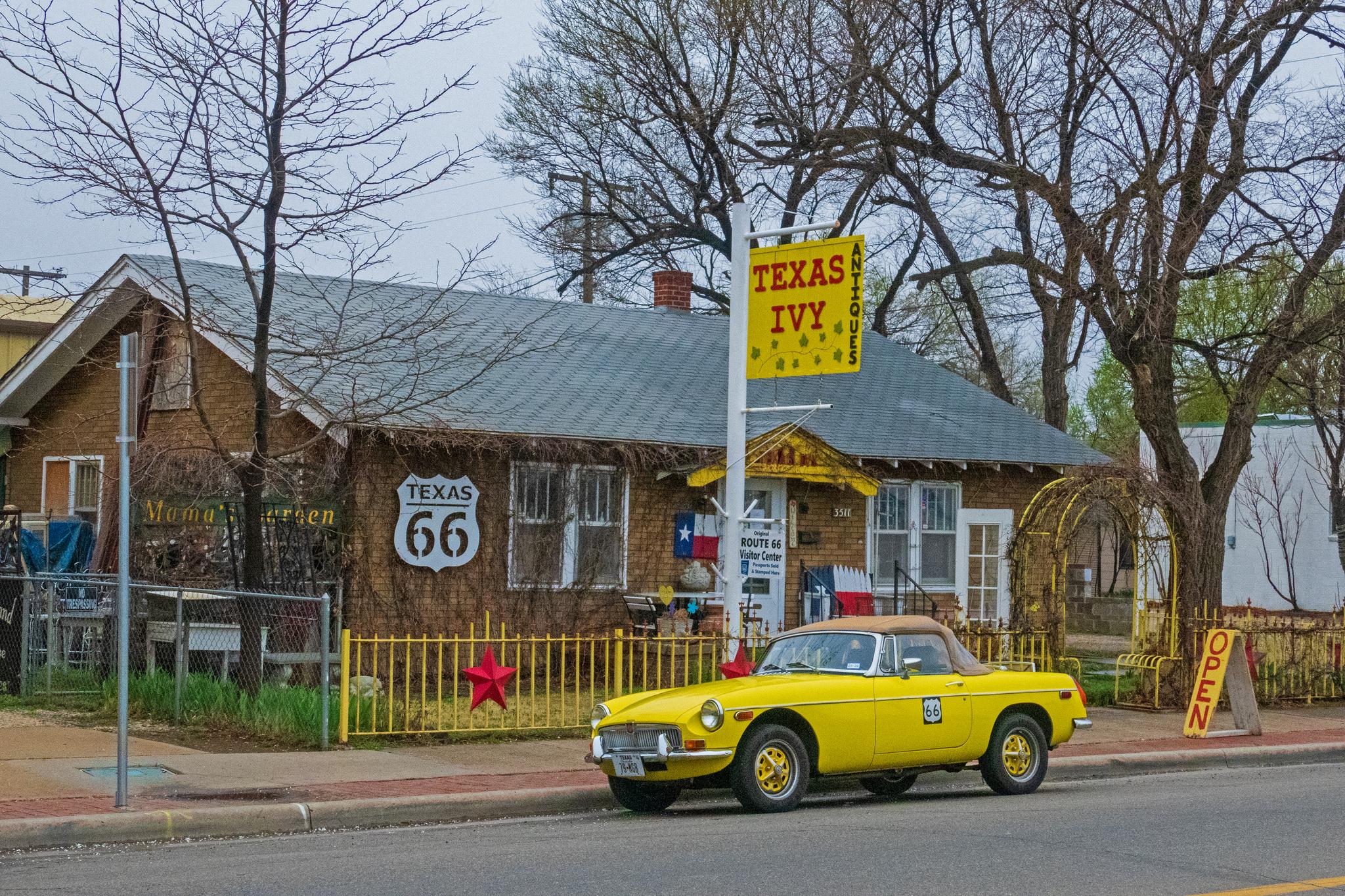 Bright yellow vintage car parked by a house with signs and bare trees.