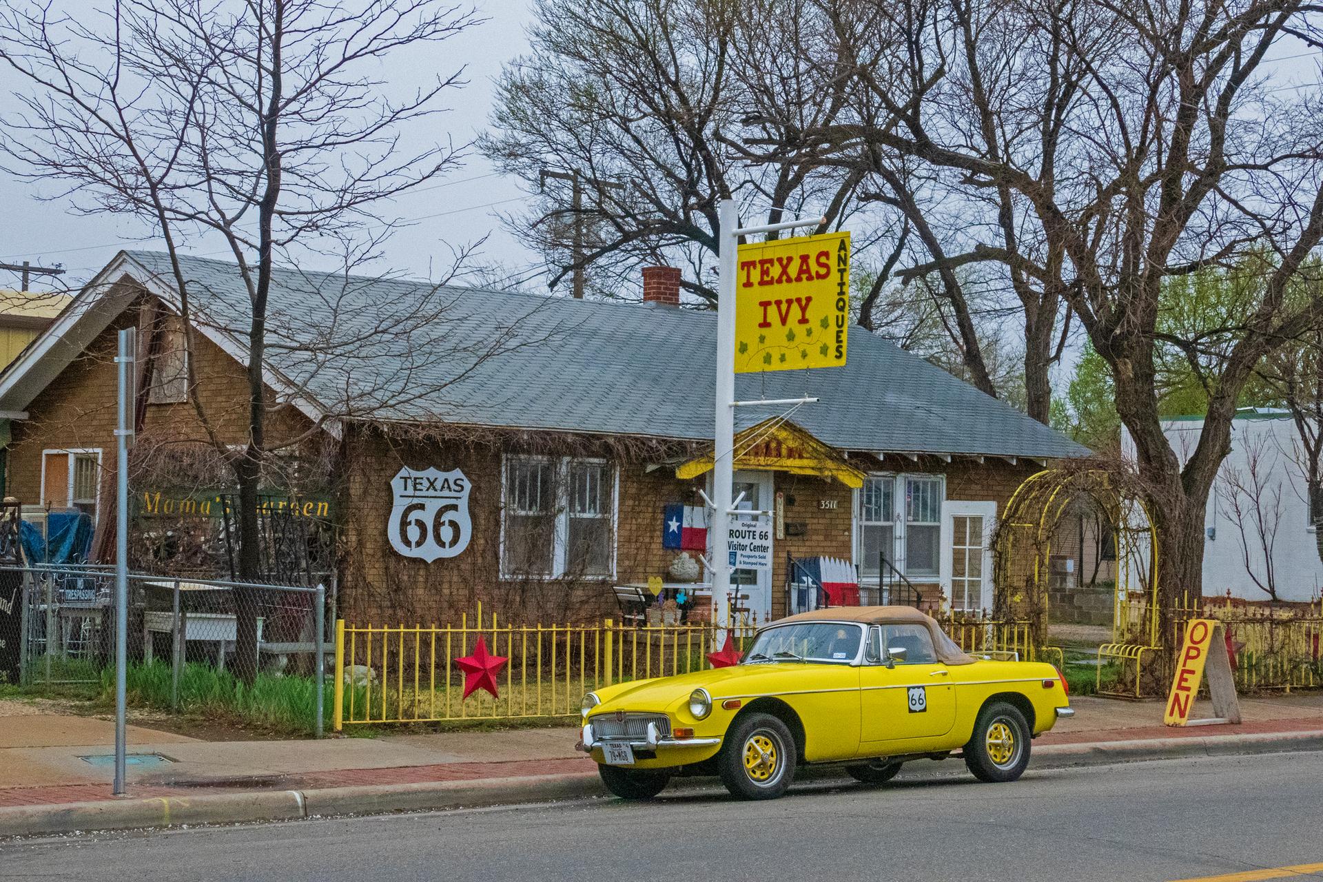 Bright yellow vintage car parked by a house with signs and bare trees.