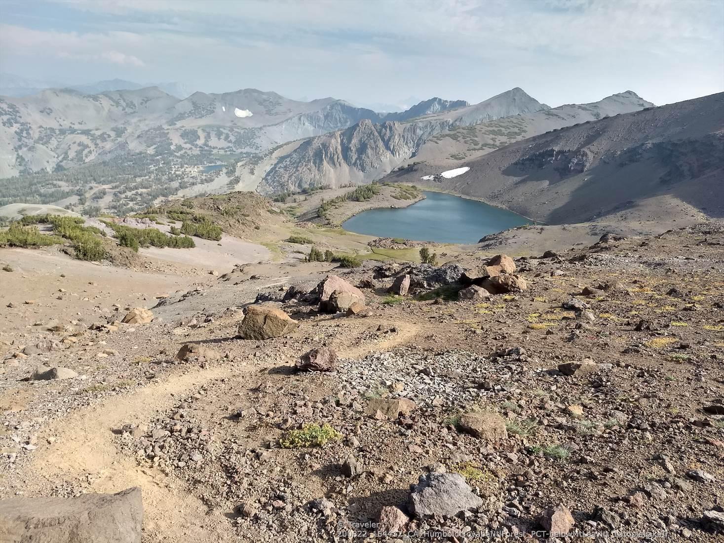 A rocky trail leads to an alpine lake surrounded by rugged mountains.