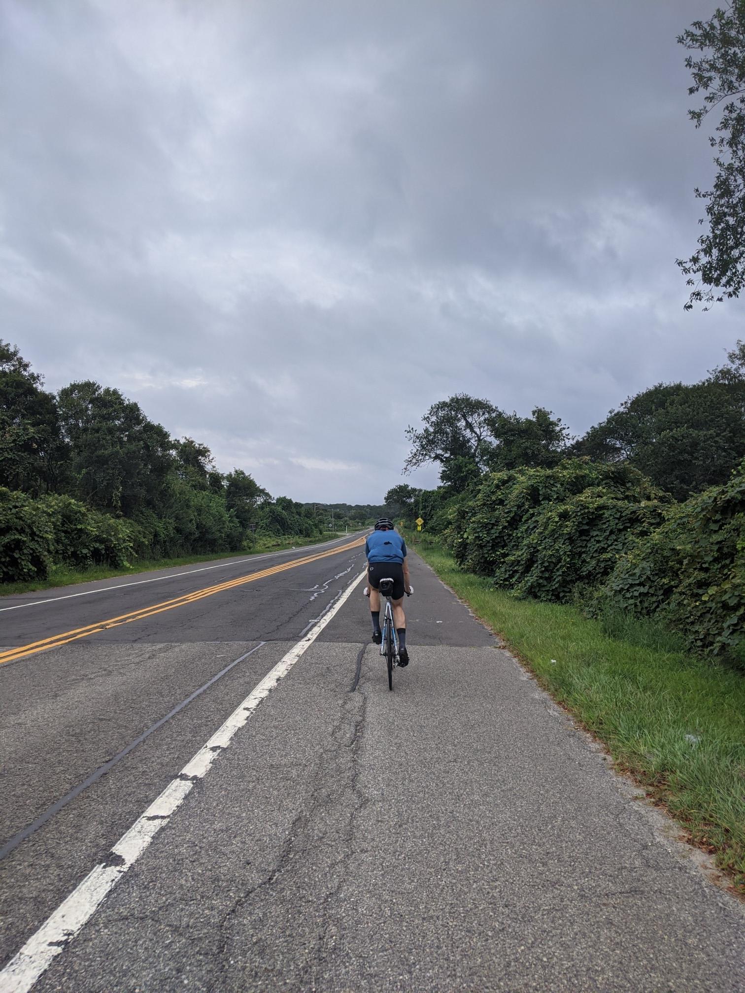 Cyclist on a paved road bordered by green trees under an overcast sky.