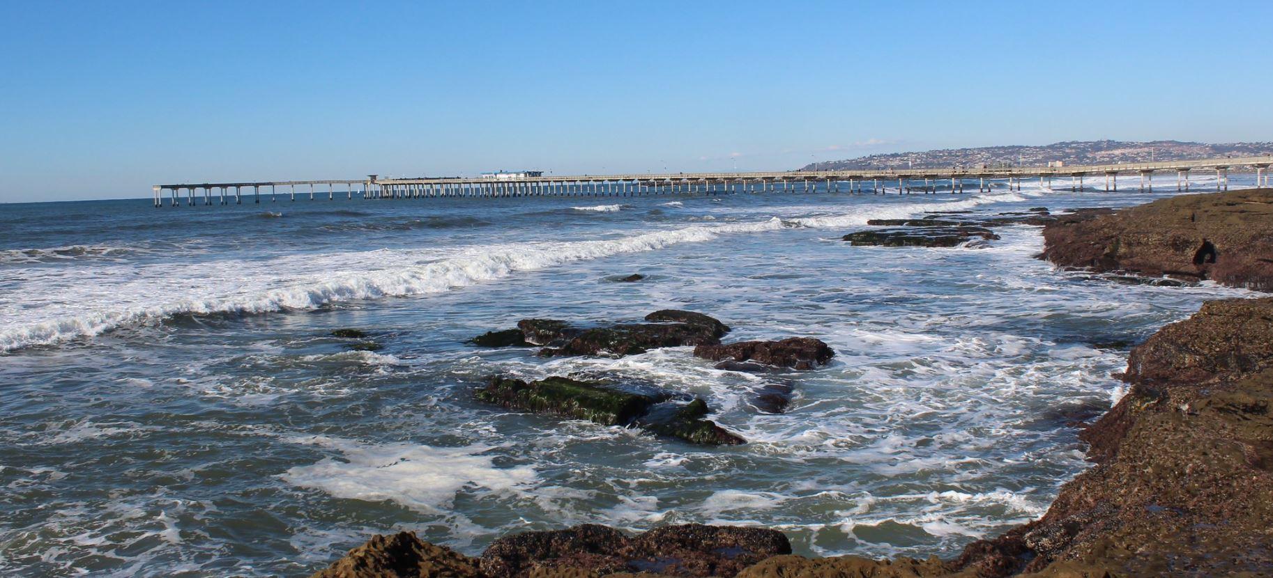 Waves crash on rocky shore with a long pier under a bright sky.