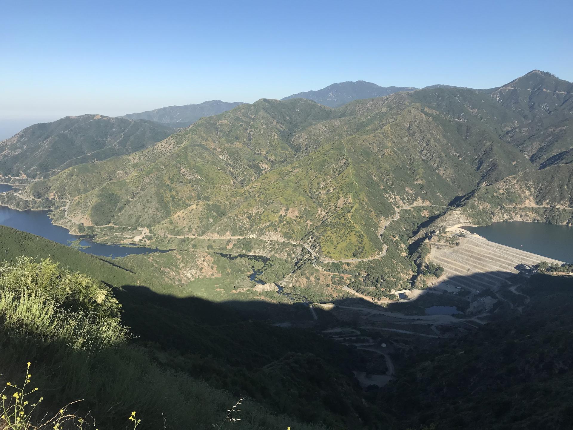 Panoramic view of green mountains, two lakes, and a large dam under clear skies.