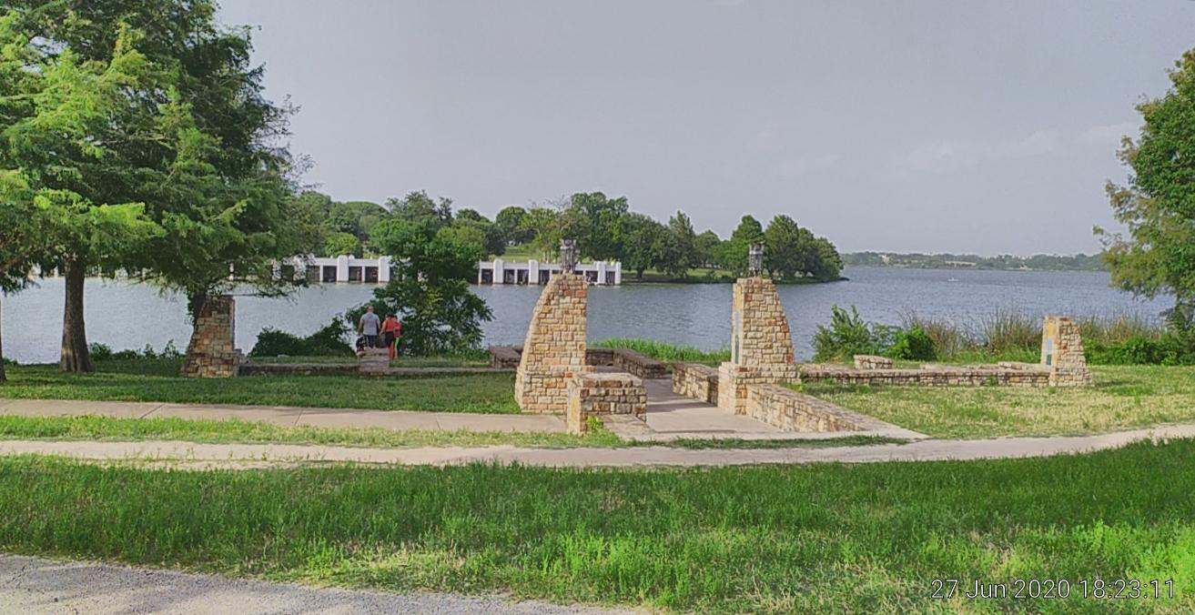 Two people sit by a calm lake with stone structures and green trees.