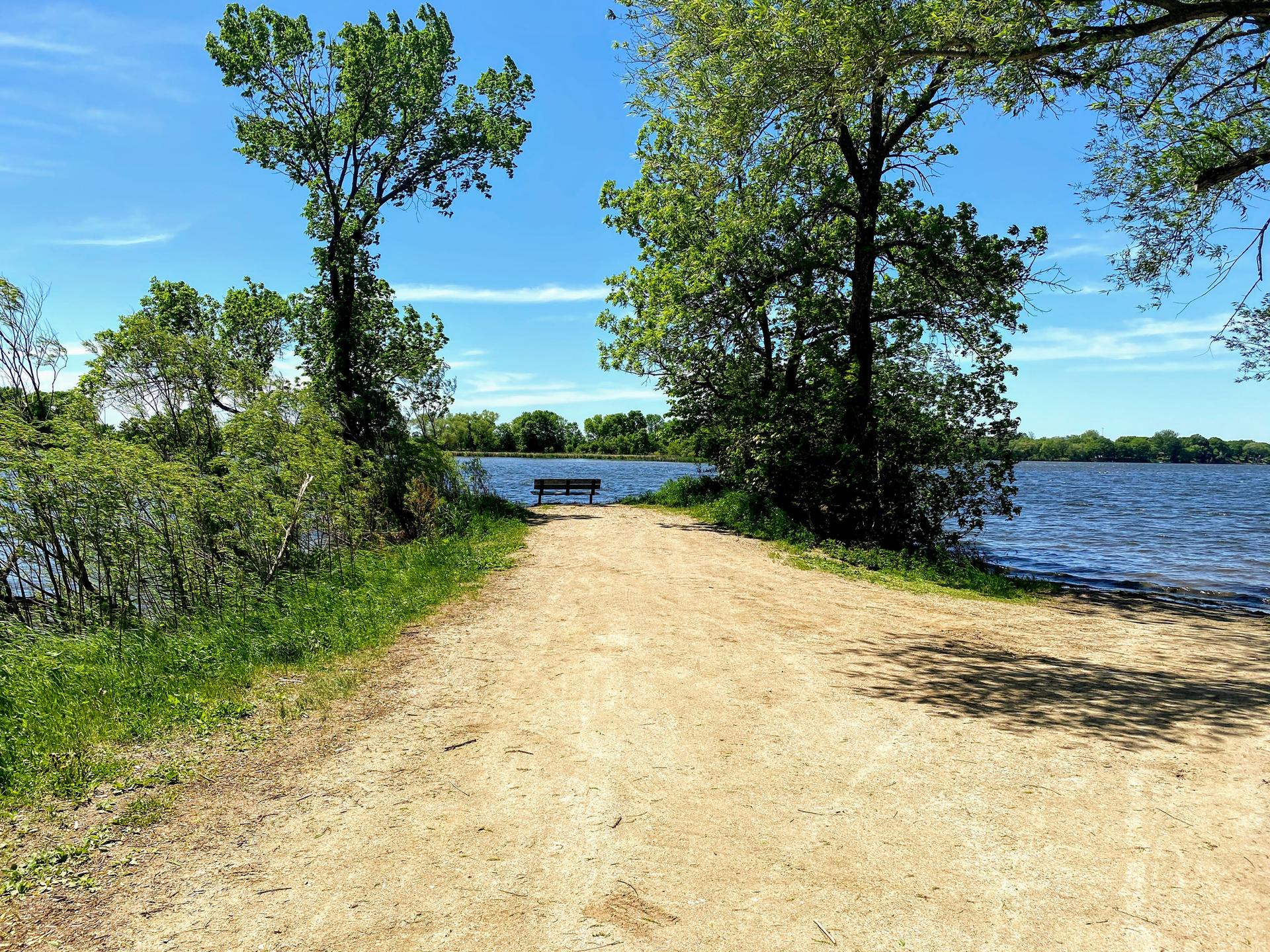 Dirt path leading to a bench overlooking a wide, sunny lake.