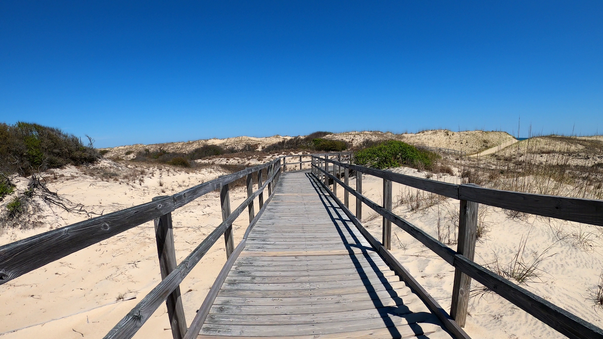 A wooden boardwalk stretches over sunny sand dunes under a clear blue sky.