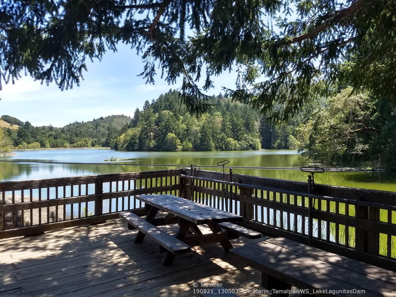 Picnic table on a shaded deck overlooking a calm lake and forest.