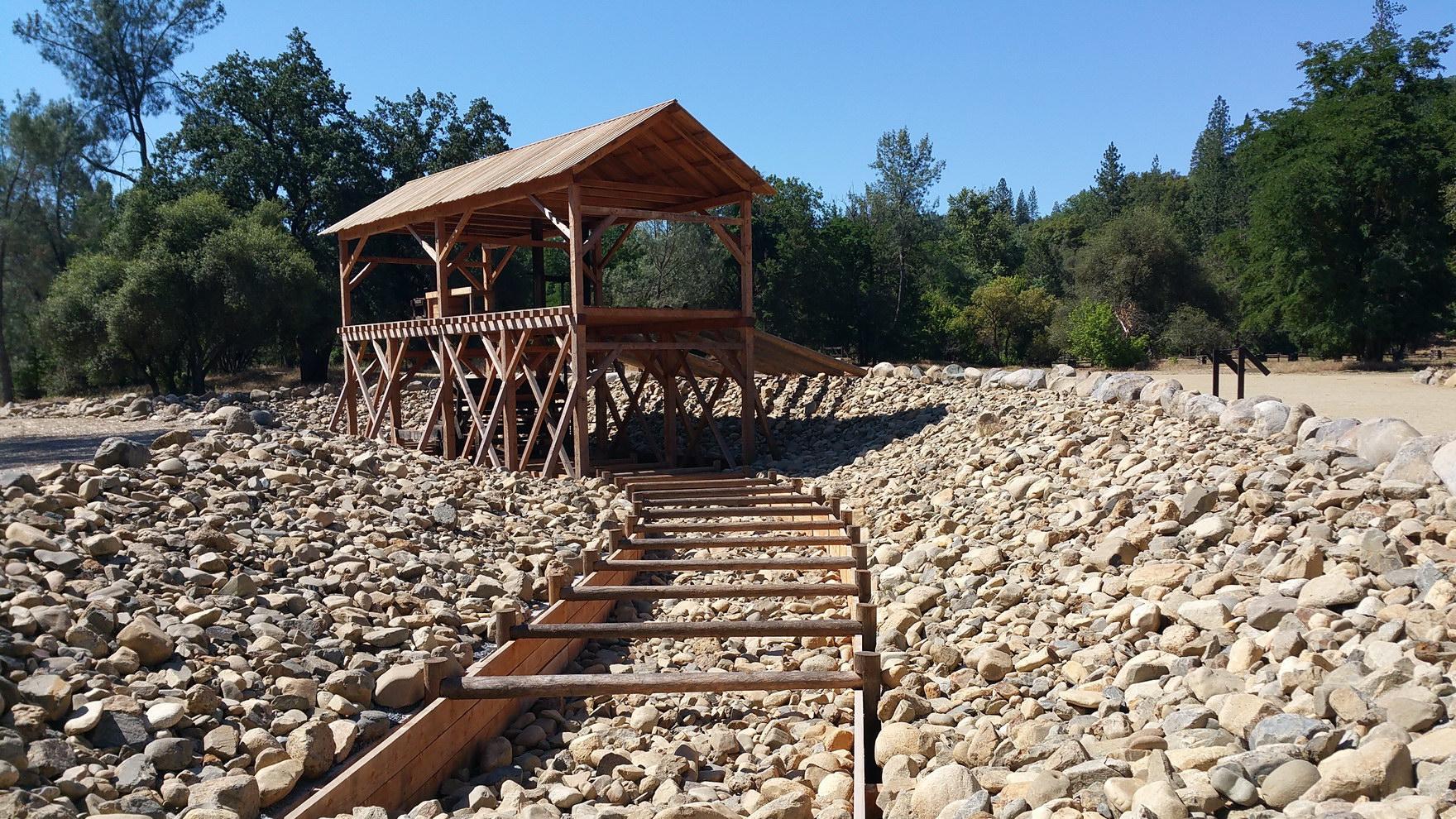 Wooden shelter and sluice box on a rocky riverbed under bright sun.