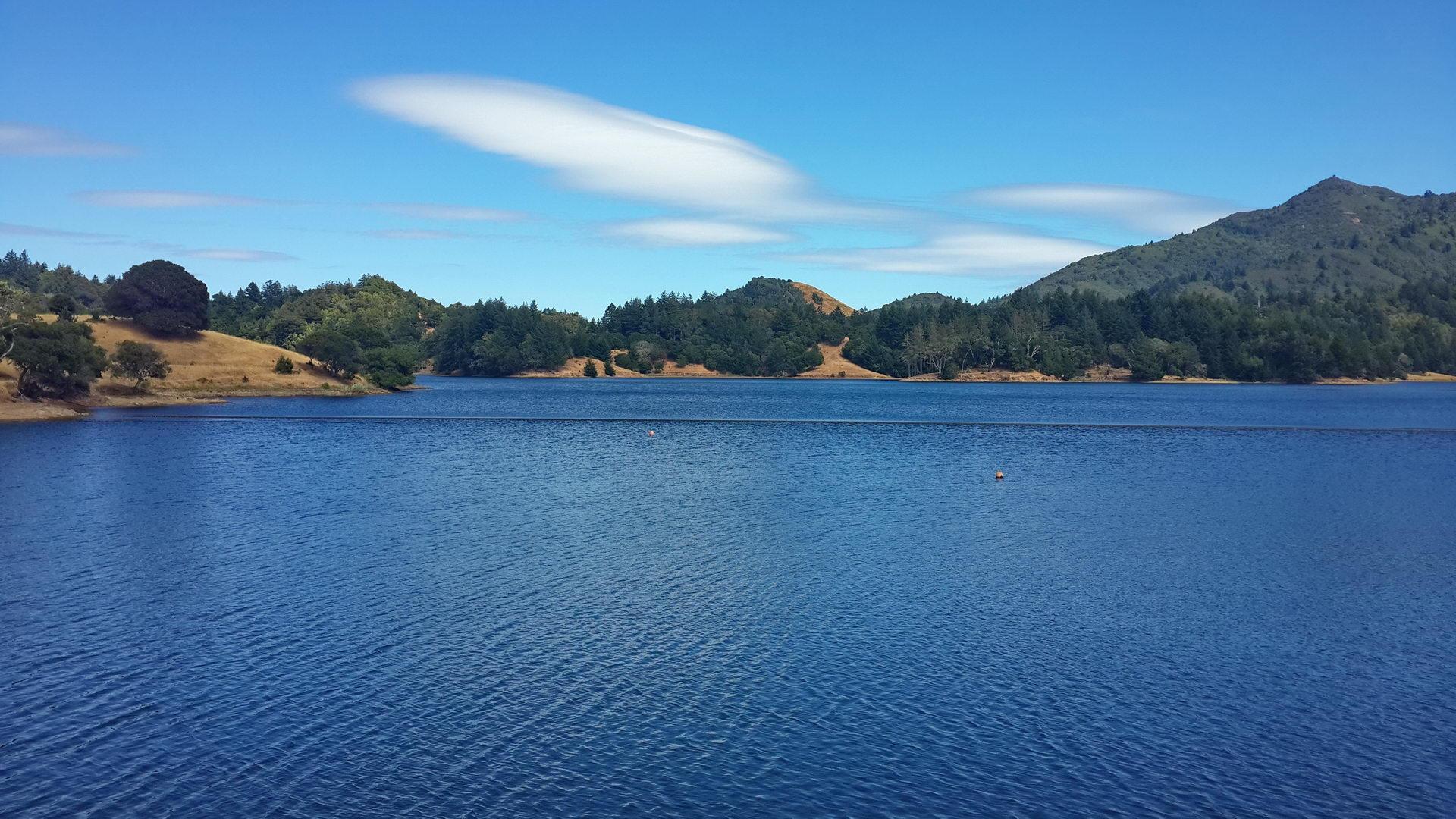 Expansive blue lake with tree-covered hills under a bright sky.