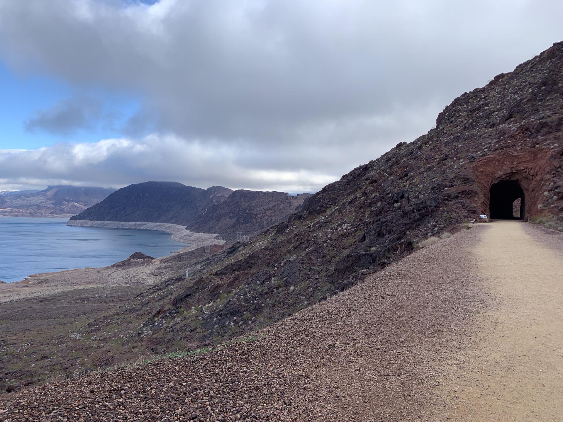 Dirt path leading to a tunnel entrance in a rocky hillside overlooking a lake.