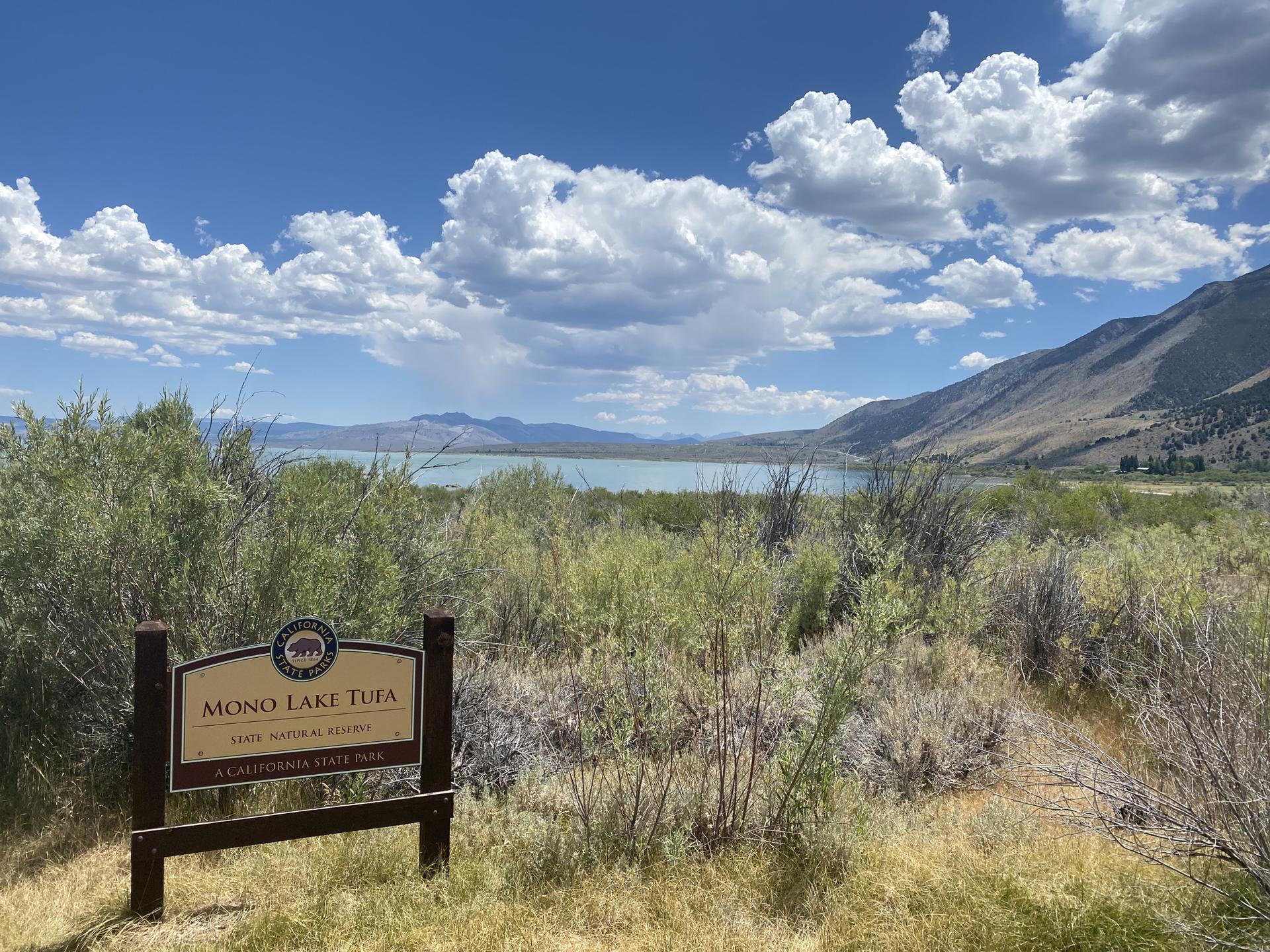 A wooden sign in a dry, brushy landscape overlooking a lake and mountains.