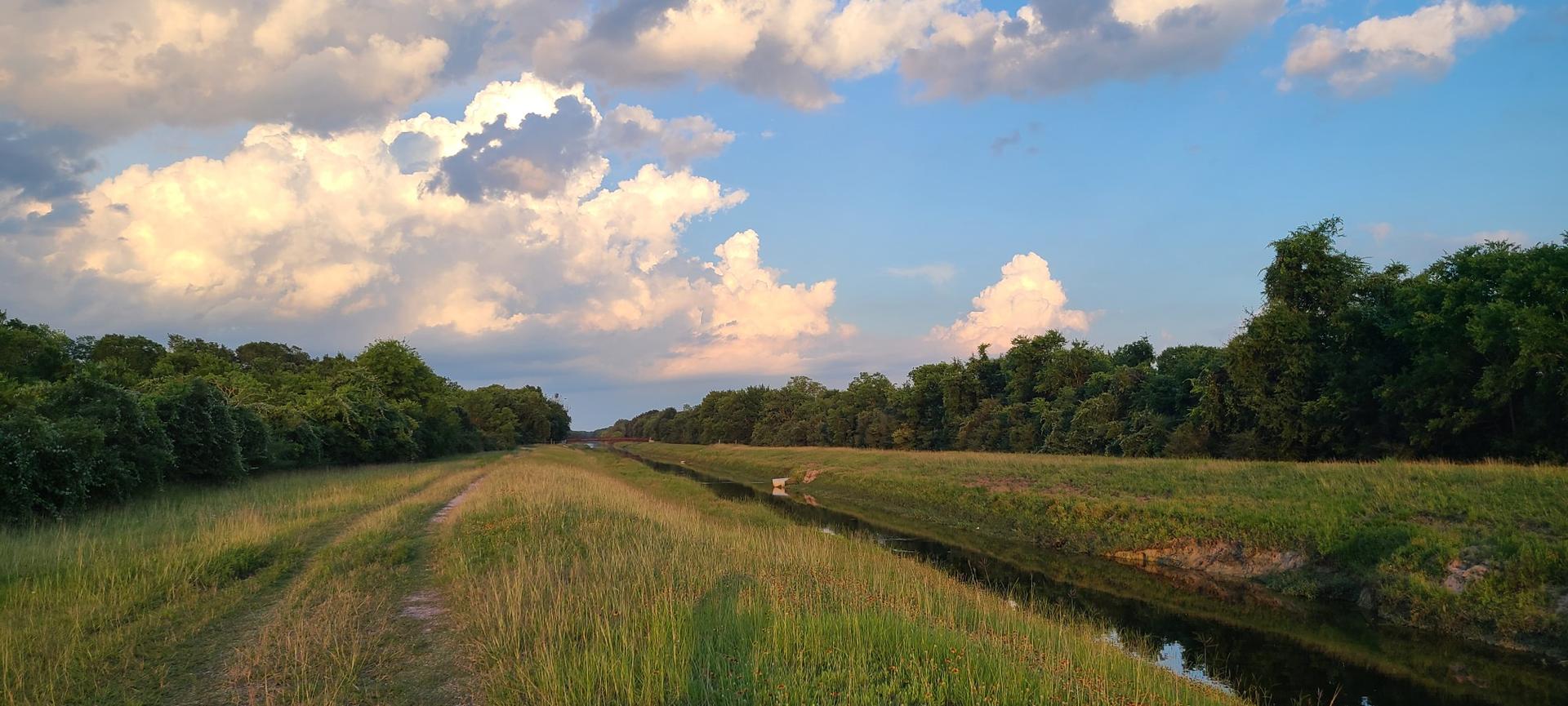 Wide grassy field with a narrow canal under a dramatic cloudy sky.