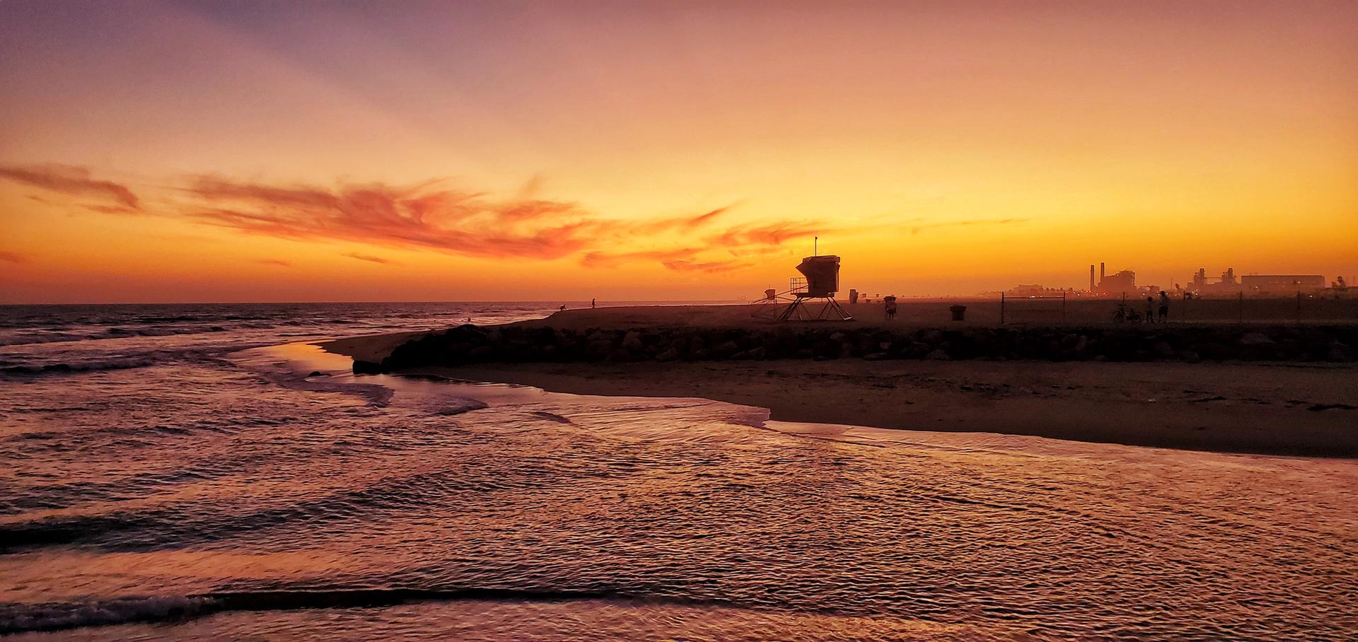 Vibrant sunset over a beach with ocean waves, a lifeguard tower, and silhouetted people.
