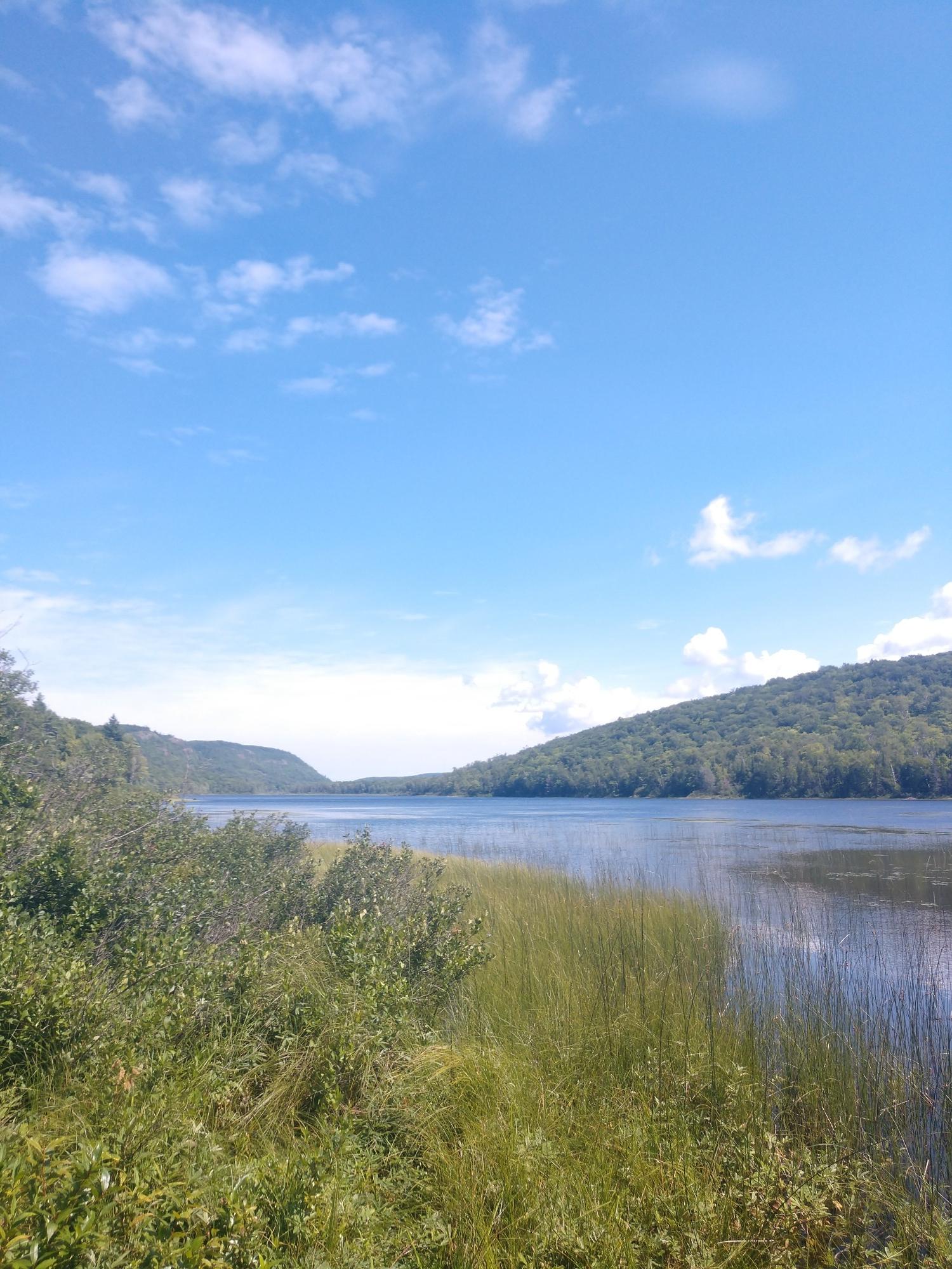 Bright daylight illuminates a calm lake bordered by green hills and tall grass.