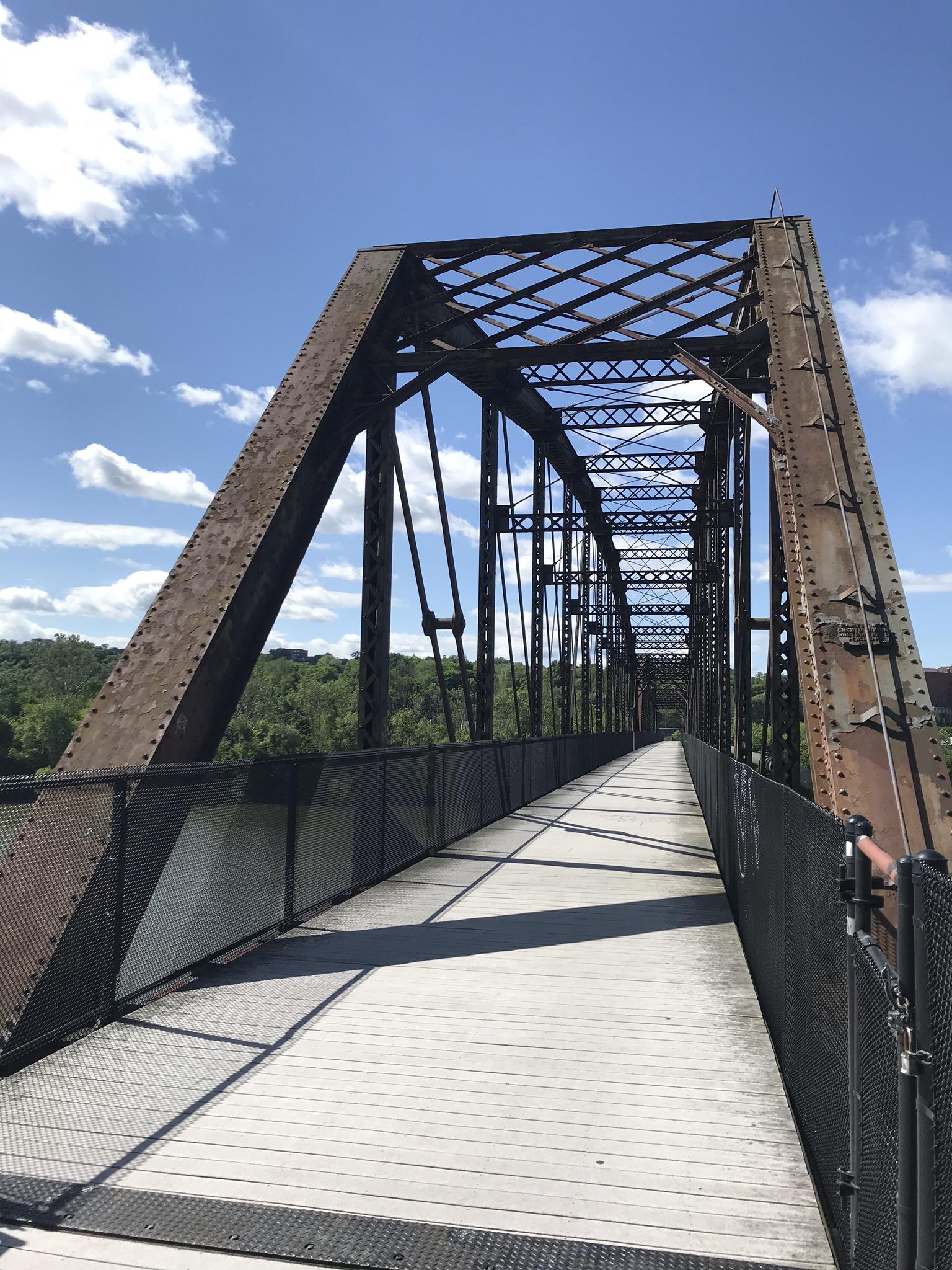 Looking down a rusty metal truss bridge walkway under a bright blue sky.