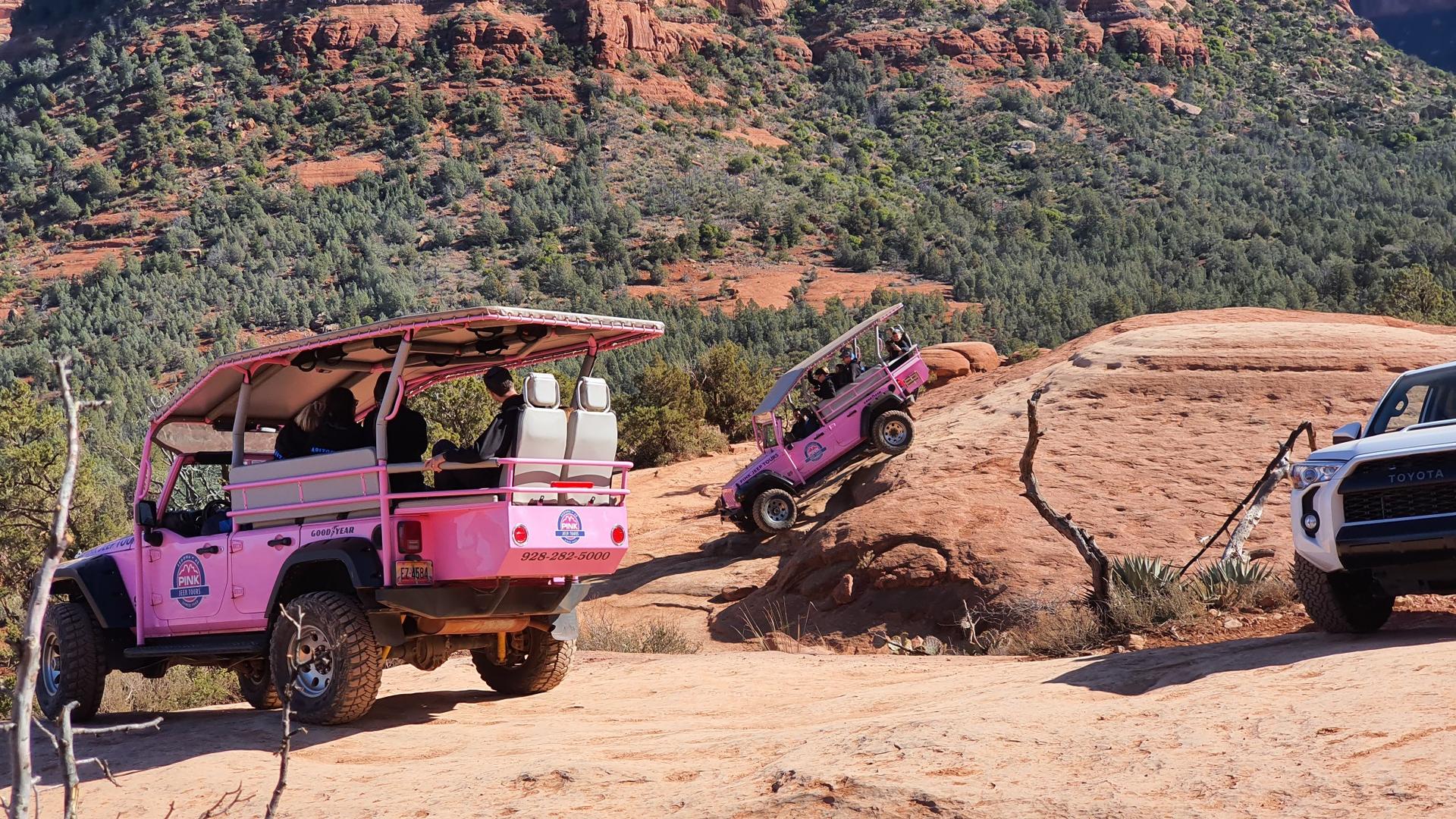 Pink off-road vehicles with passengers navigating a sunny, rocky desert terrain.