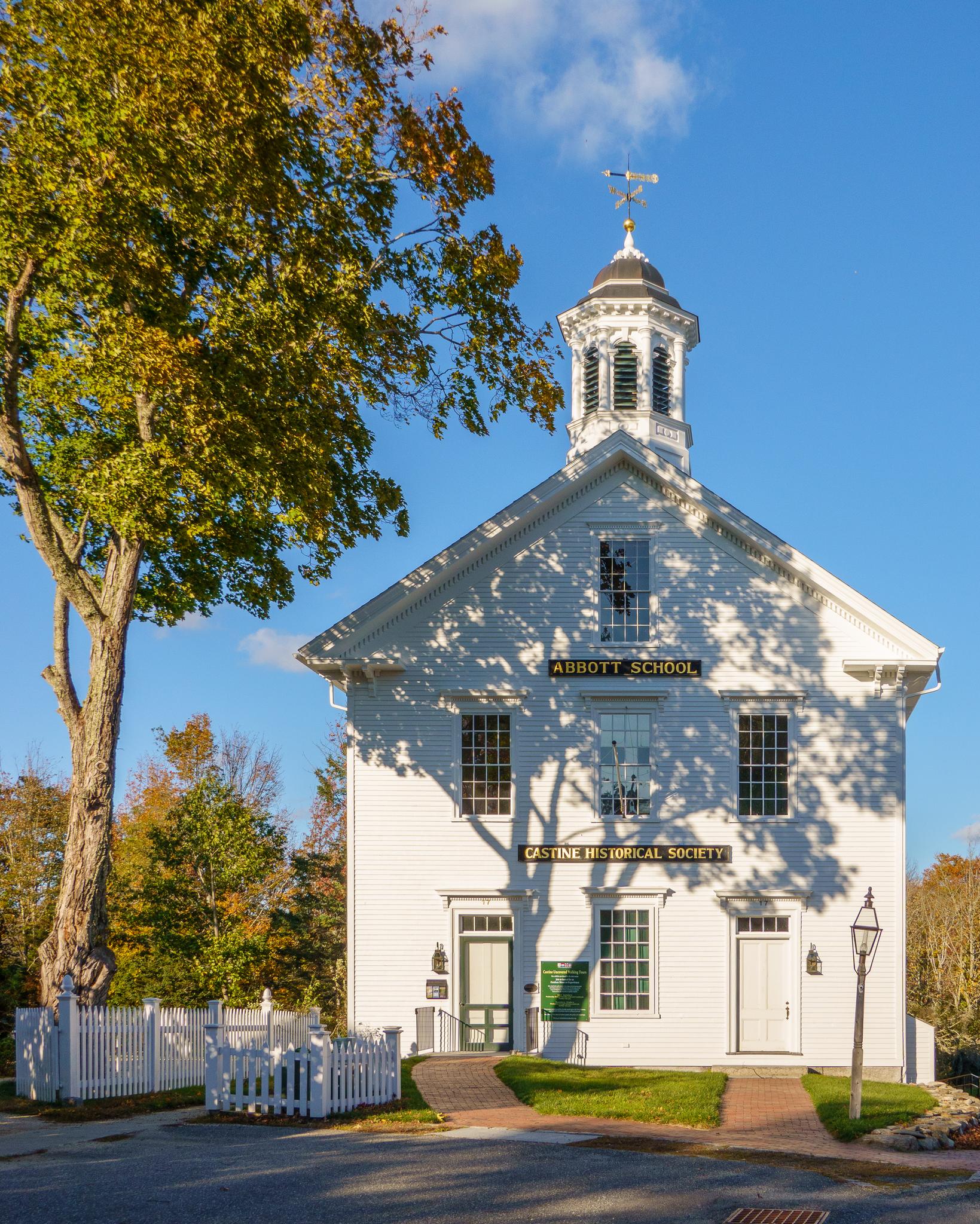 A white historical building with a cupola, large tree, and white picket fence.