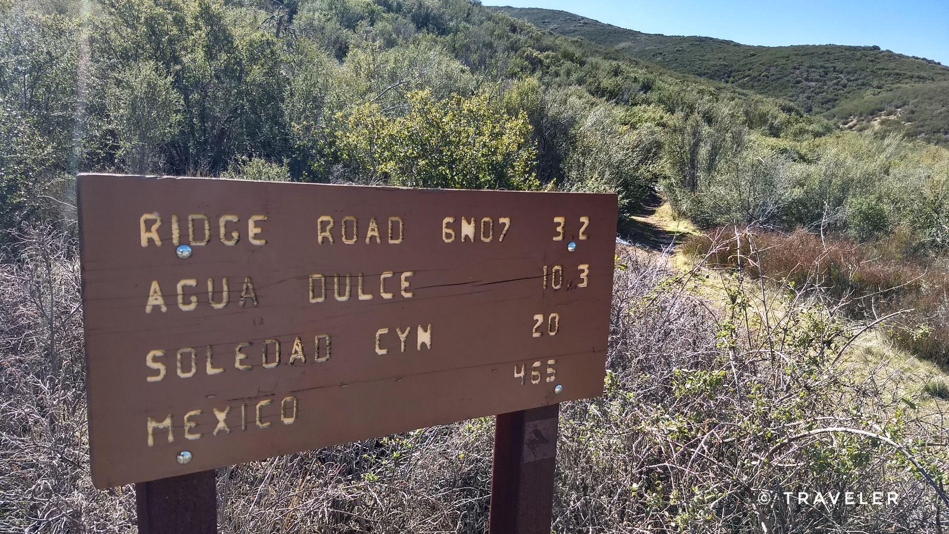Close-up of a brown wooden trail sign in a sunny, brushy landscape.