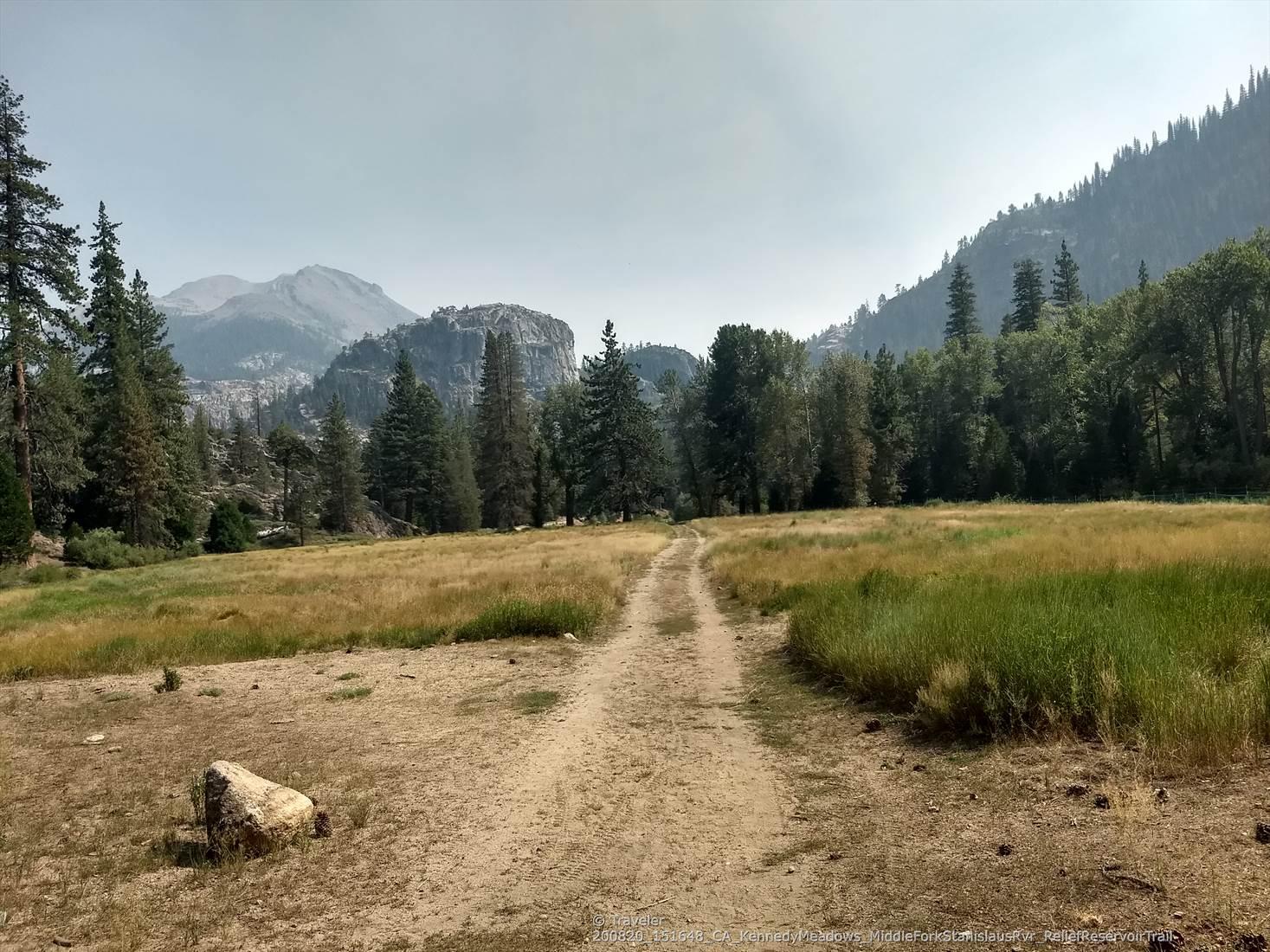 A dirt path winds through a hazy meadow towards distant mountains.