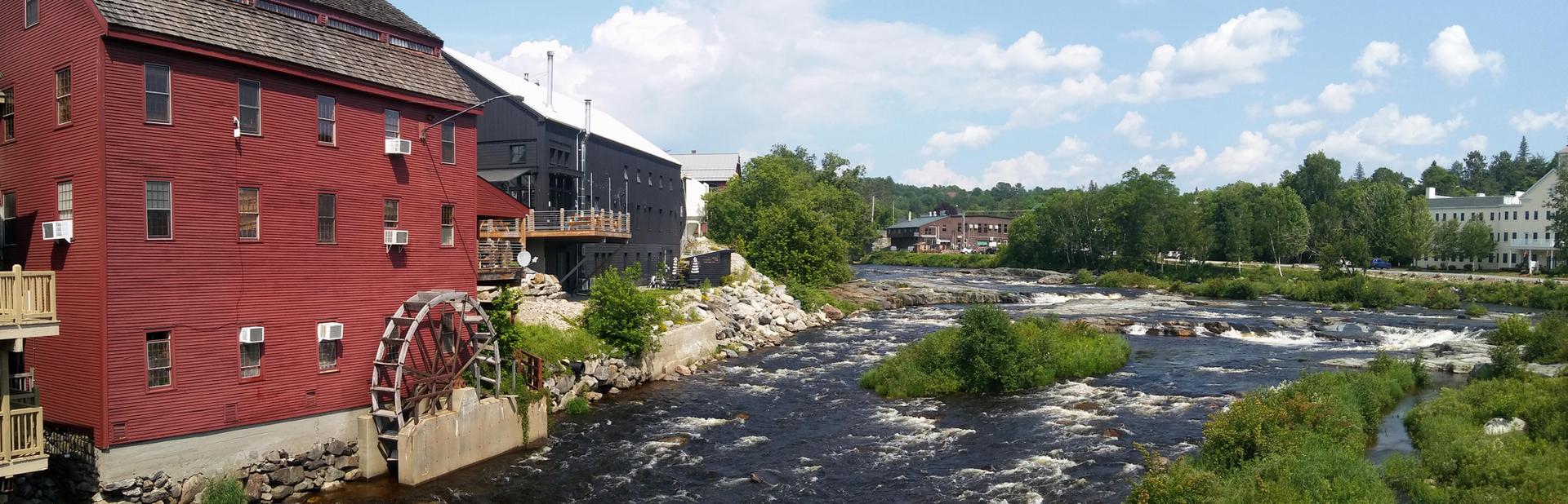 Red building with water wheel beside a river with rapids and distant town.