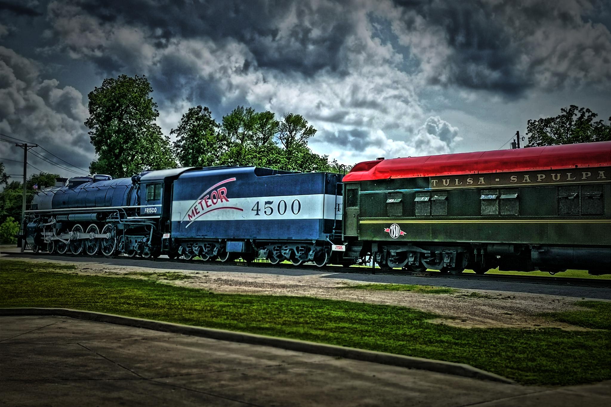 Two vintage train cars, blue and green, parked under a dramatic cloudy sky.