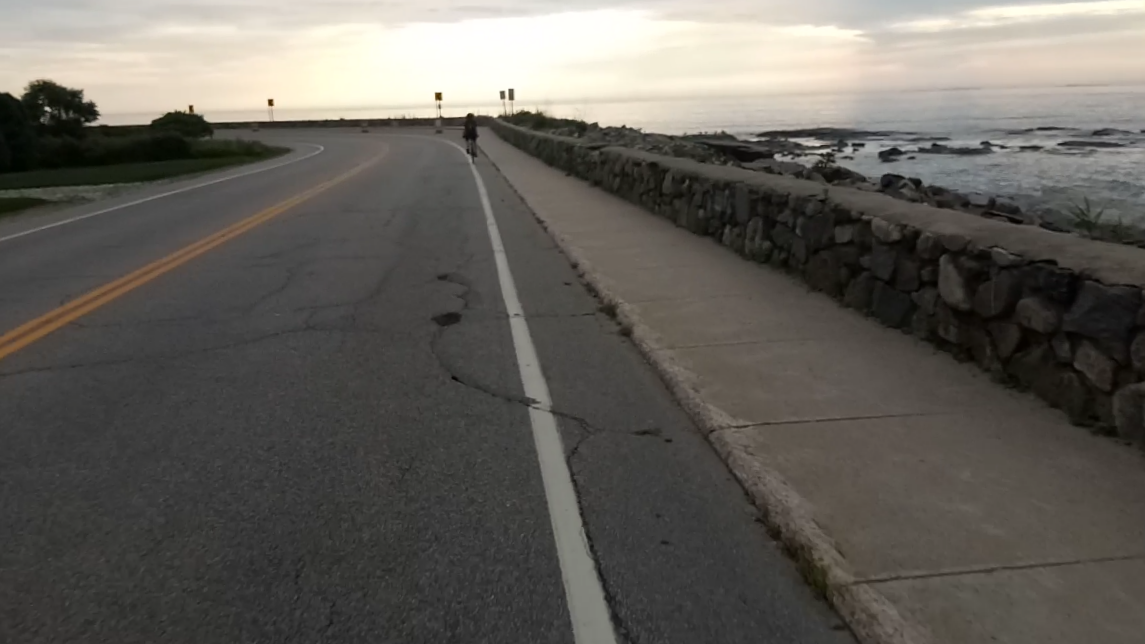 A person cycles on a coastal road next to a stone wall at dusk. - East Coast Greenway: Portsmouth Coastal Loop