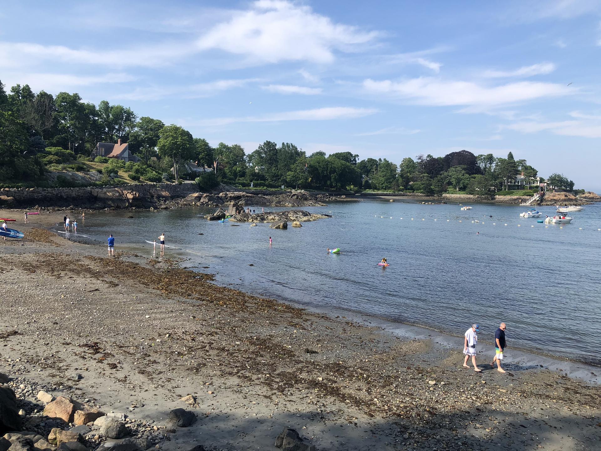 People on a rocky beach and in calm water with boats under sunny sky.