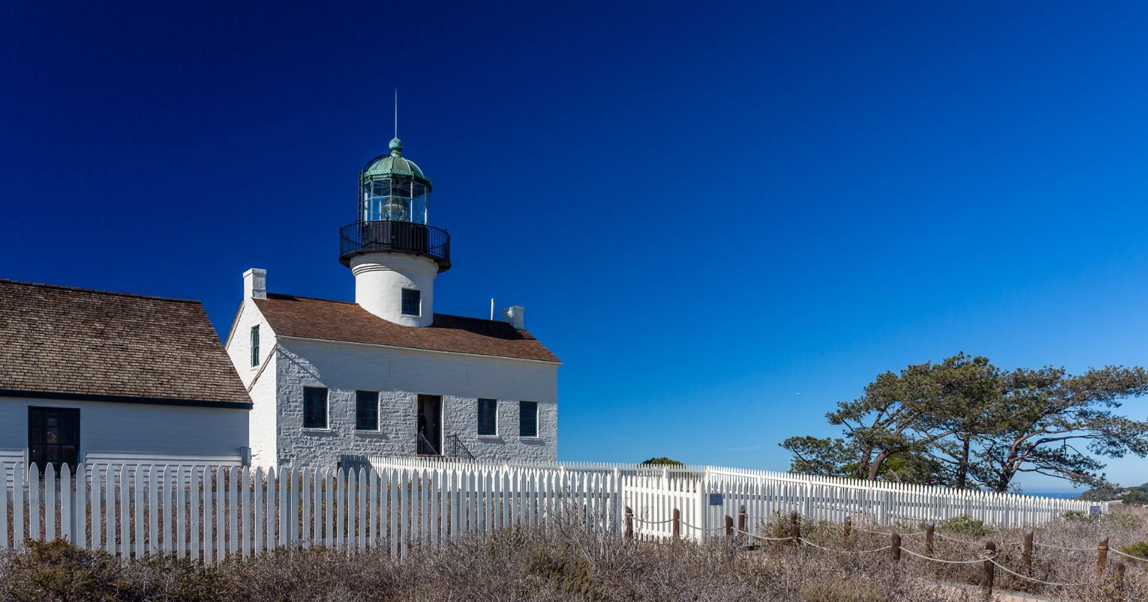 White lighthouse with a green dome, white picket fence, clear blue sky. - Old Point Loma Lighthouse – Point Loma loop from Cabrillo National Monument