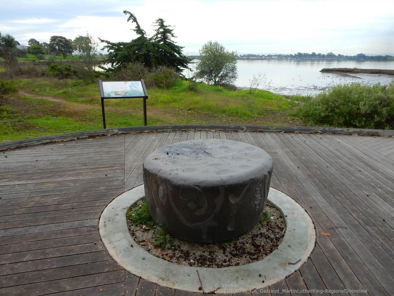 Stone sculpture on a wooden deck overlooking a calm body of water.