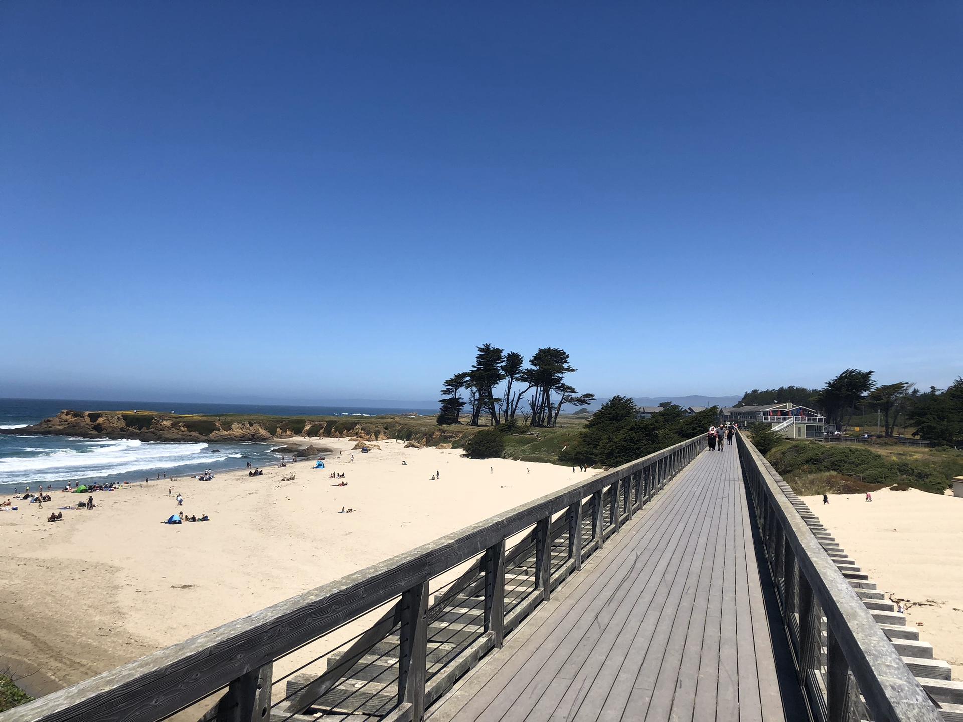 Long wooden boardwalk beside a sunny beach with people and ocean waves.