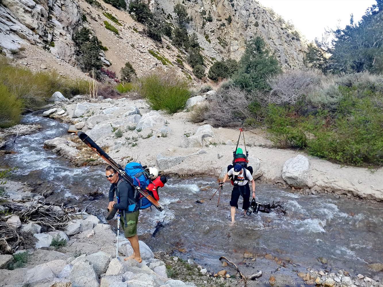 Two hikers with skis and backpacks wade through a rocky mountain stream.