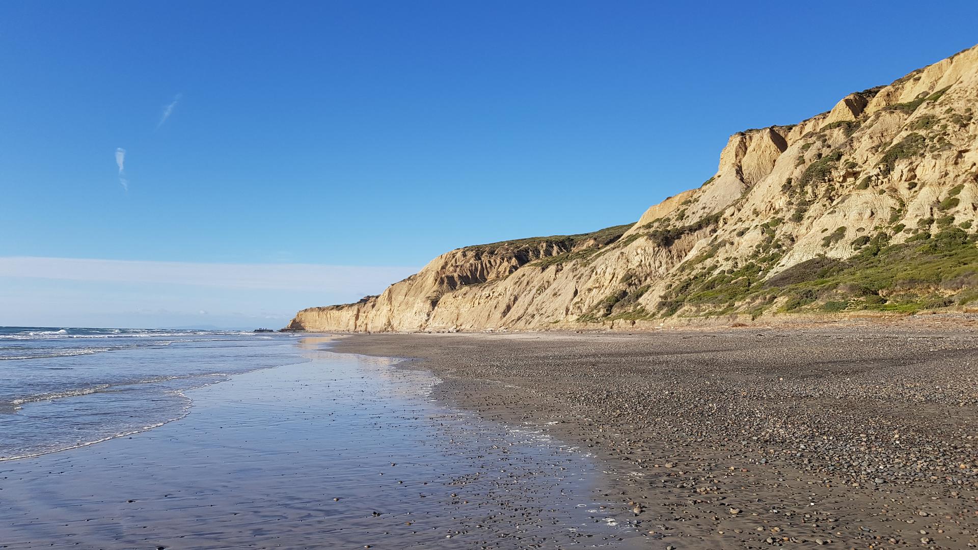 Sunny beach with calm ocean waves and towering light-colored cliffs.