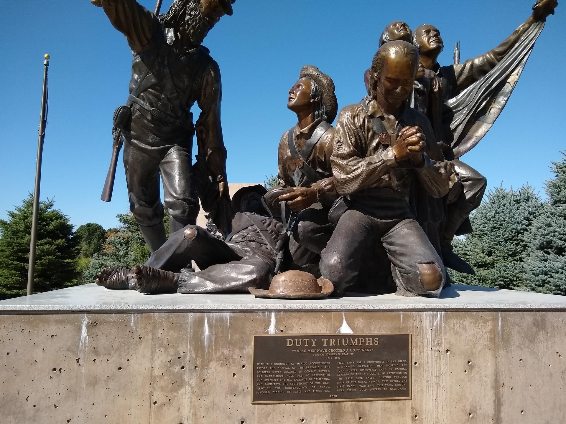 A bronze statue depicts multiple figures, some kneeling, under a bright blue sky.