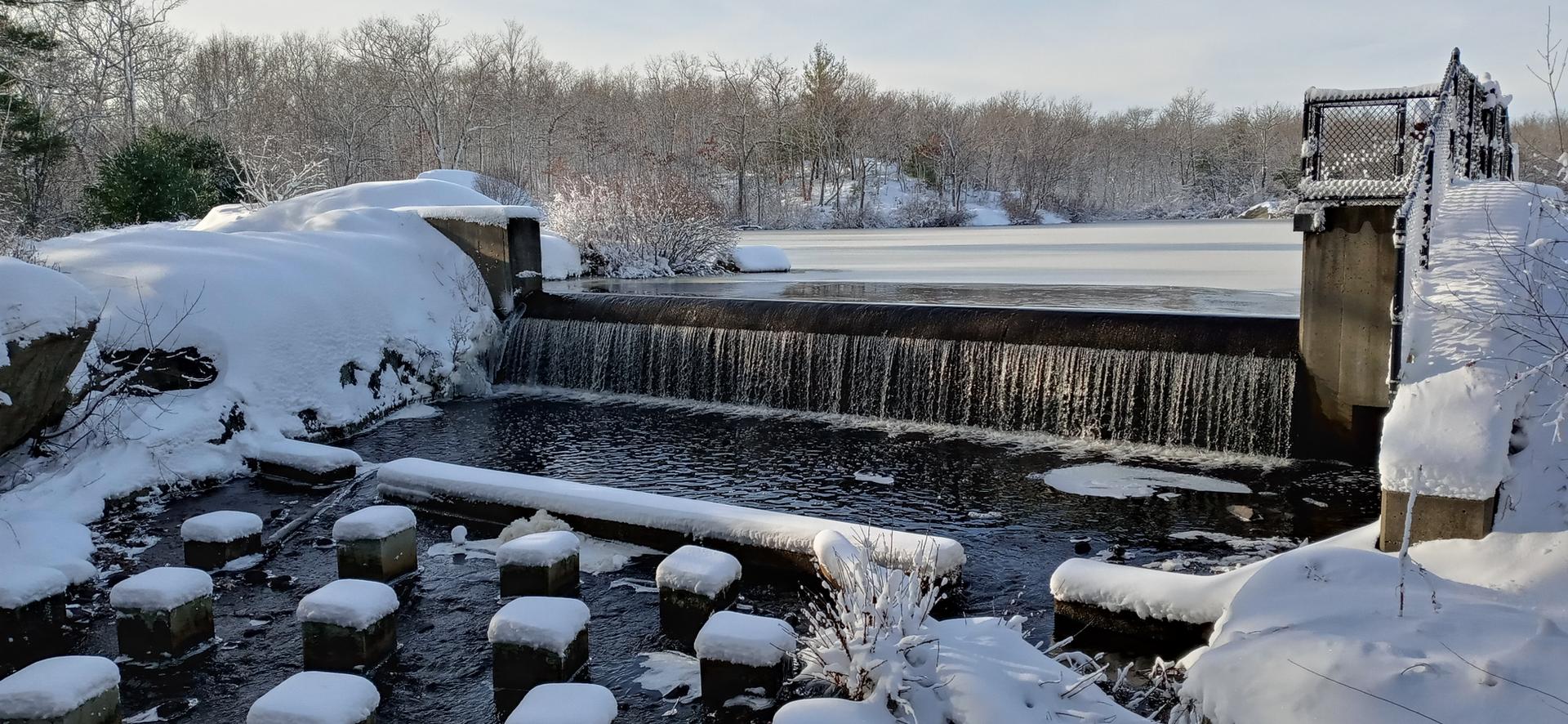 Snow-covered dam with a small waterfall, surrounded by winter landscape.