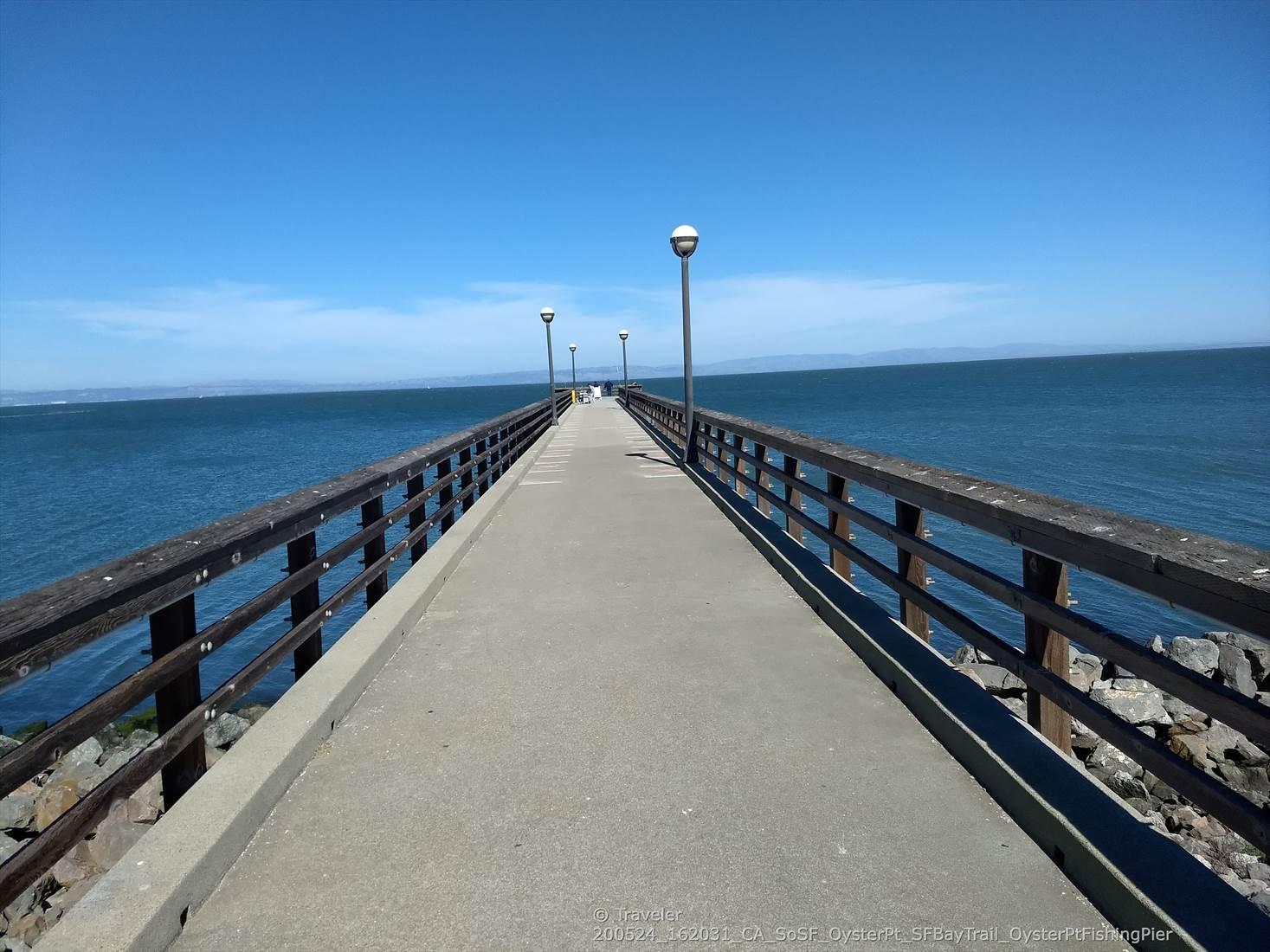Long concrete pier with railings and lampposts extending into vast blue water.