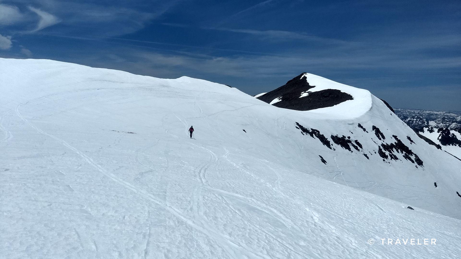 A lone person walks across a vast, snow-covered mountain slope under a blue sky.