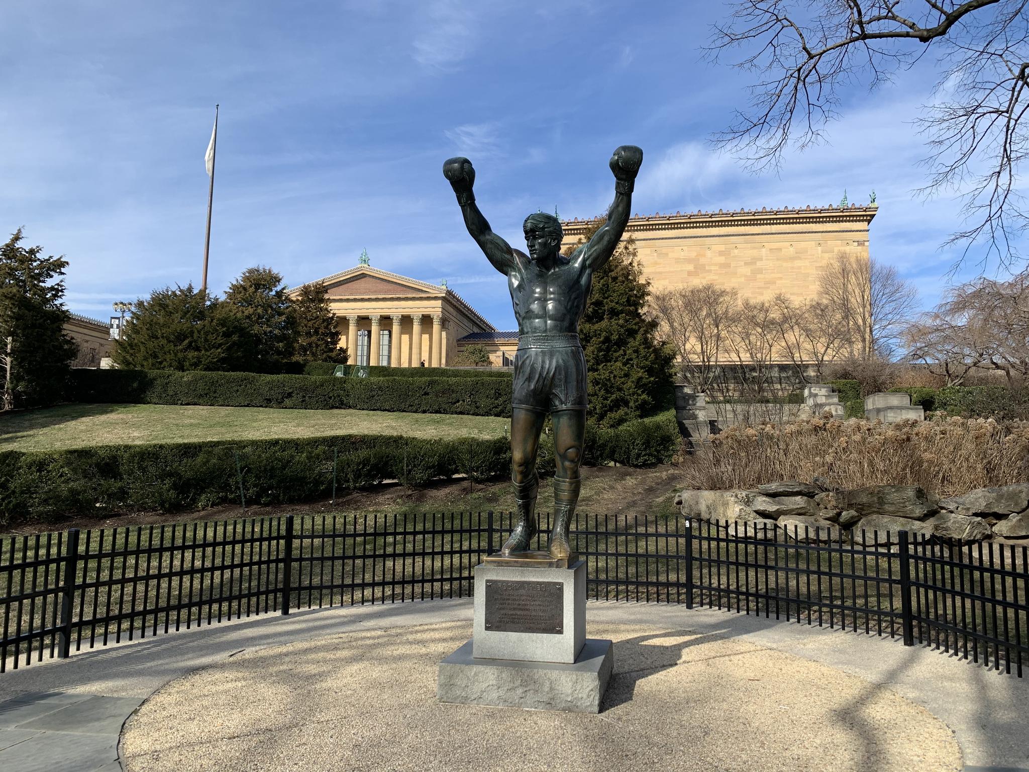 Bronze statue of a triumphant man with raised arms in a park setting.