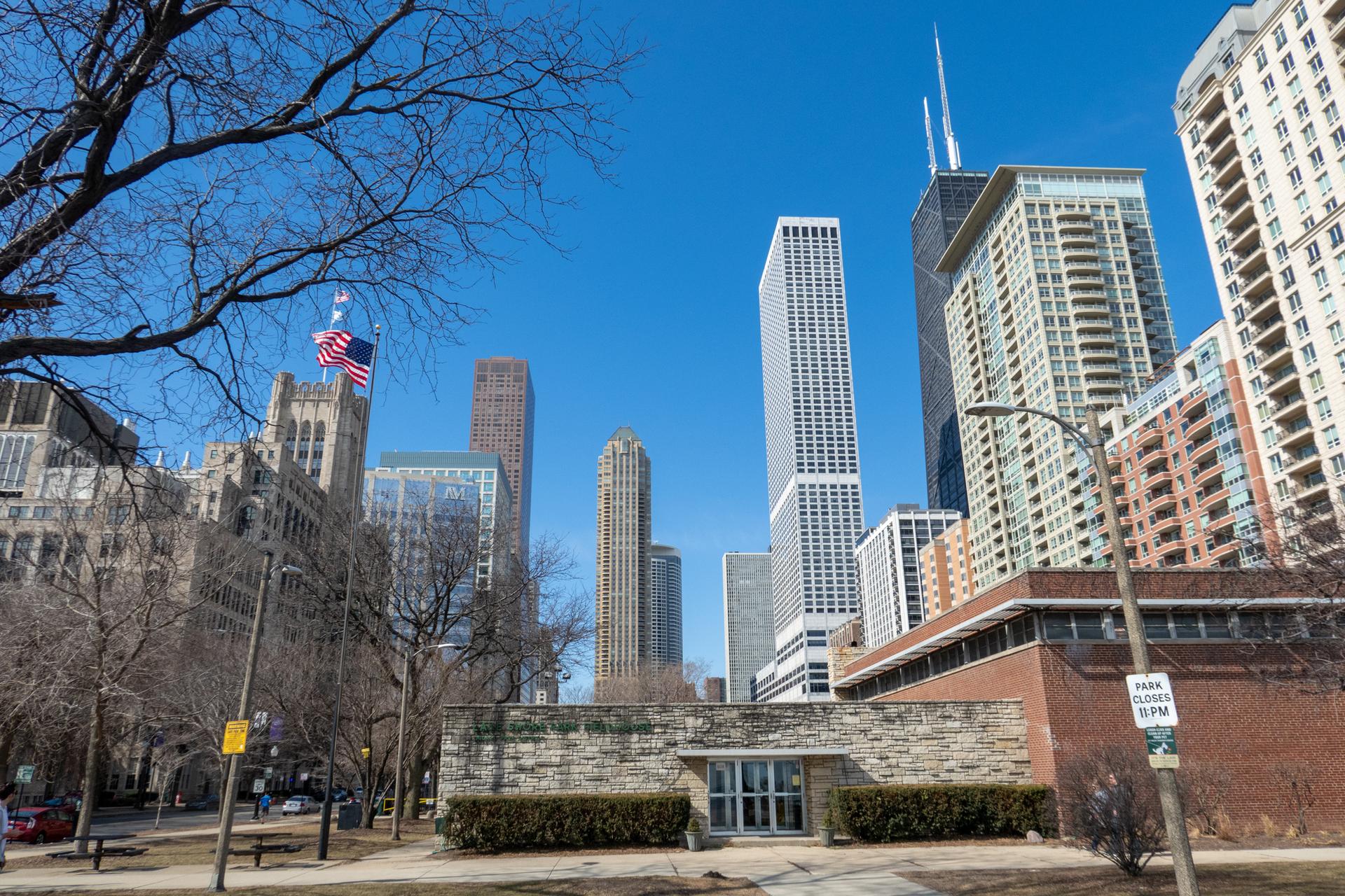 Tall city buildings under a clear blue sky, with bare trees in foreground.