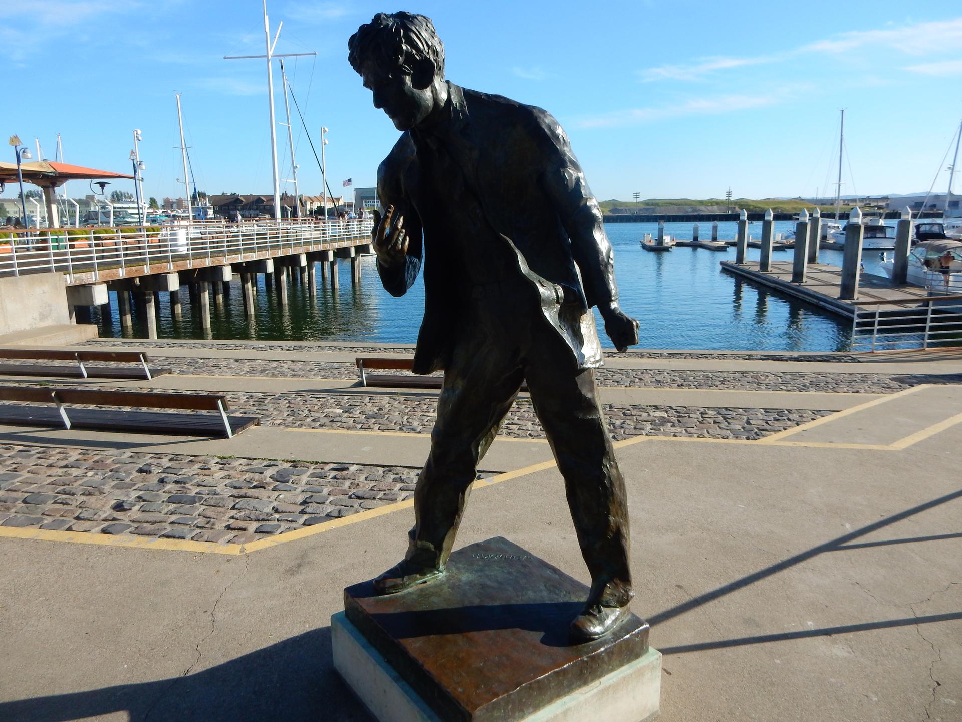 Bronze statue of a man on a paved area next to a marina with boats. Sunny.
