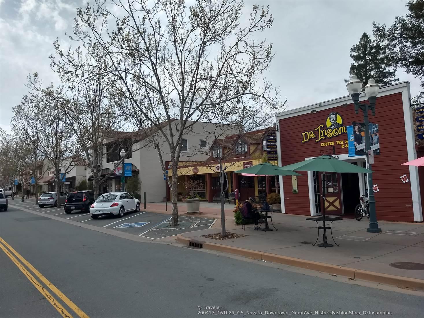 Urban street with shops, bare trees, parked cars, and people dining outdoors.