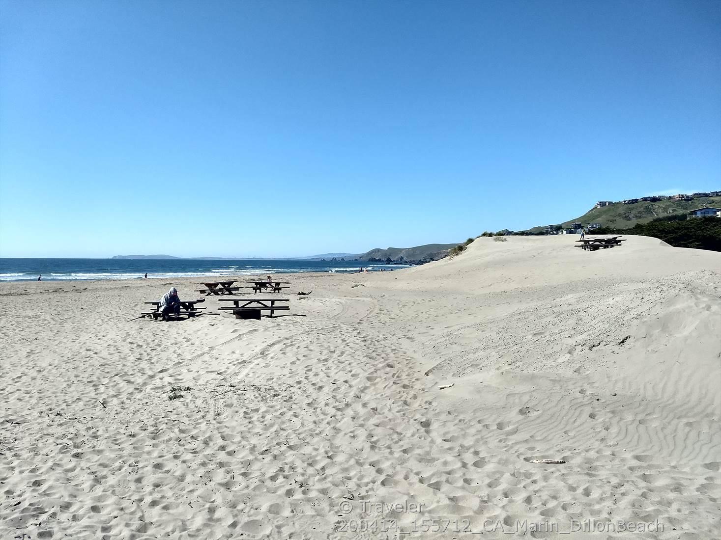 A wide sandy beach with picnic tables, ocean, and distant hills.