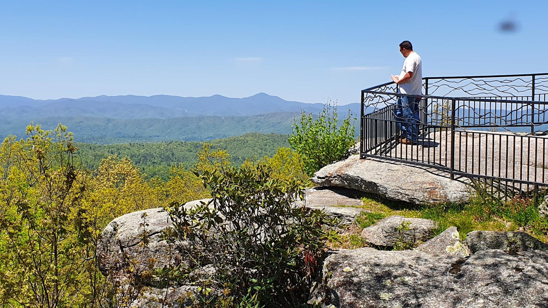 Man on a sunny mountain overlook, gazing at distant peaks.