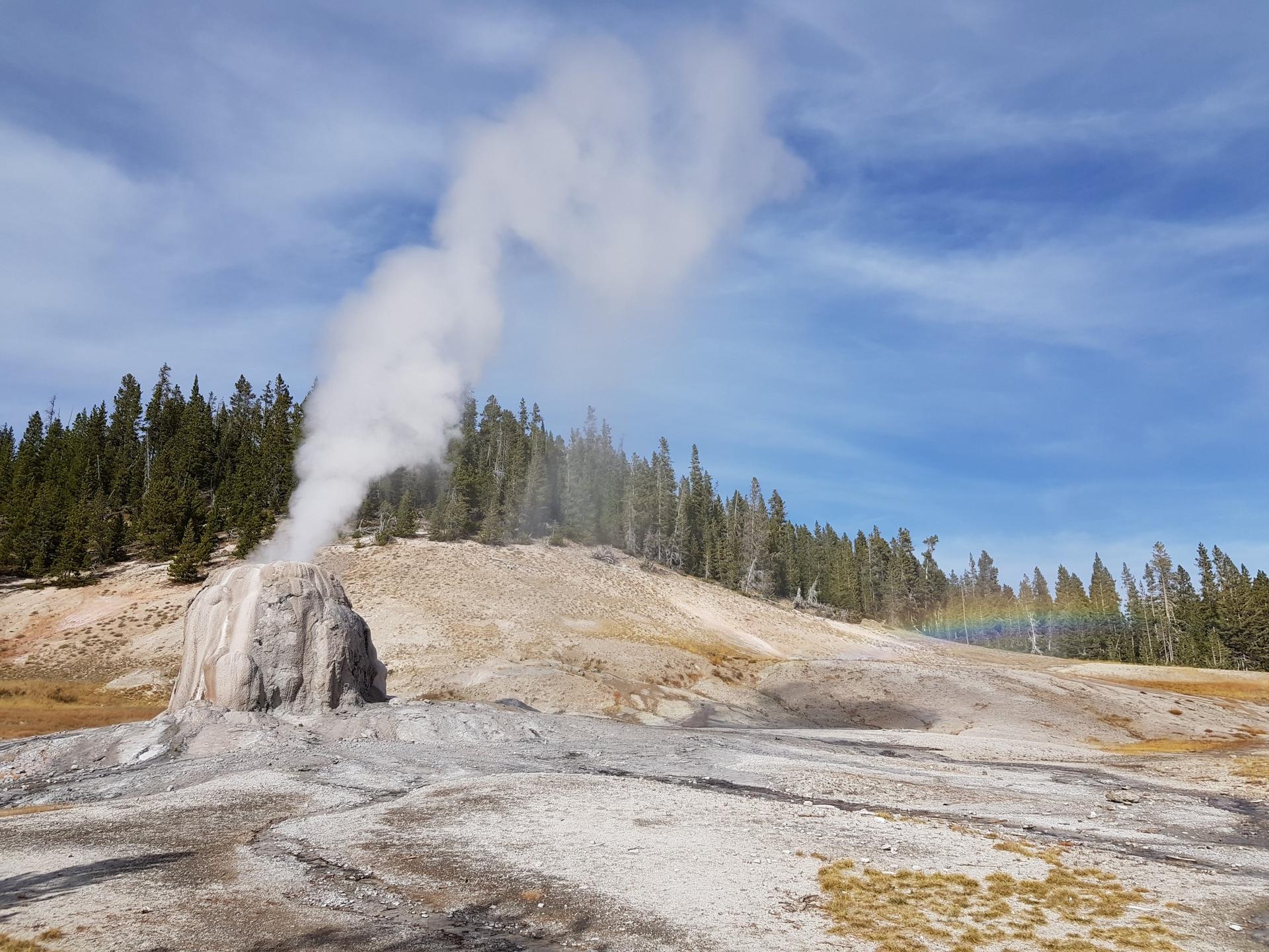 Geyser erupting steam into a blue sky with a faint rainbow.