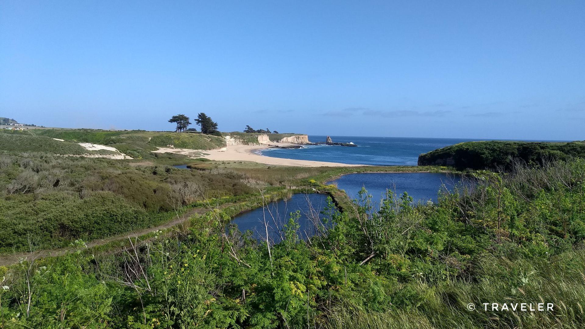 Panoramic view of green coastal hills, ponds, a sandy beach, and ocean.