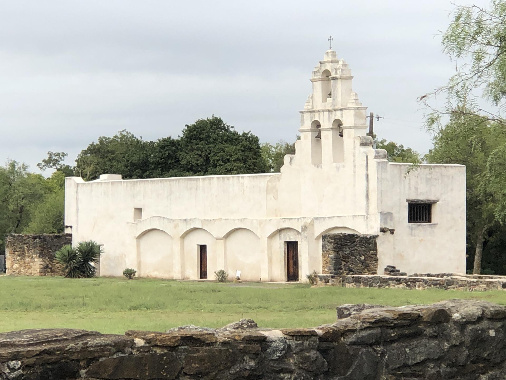 White stucco building with bell tower and arched entrances on a cloudy day.