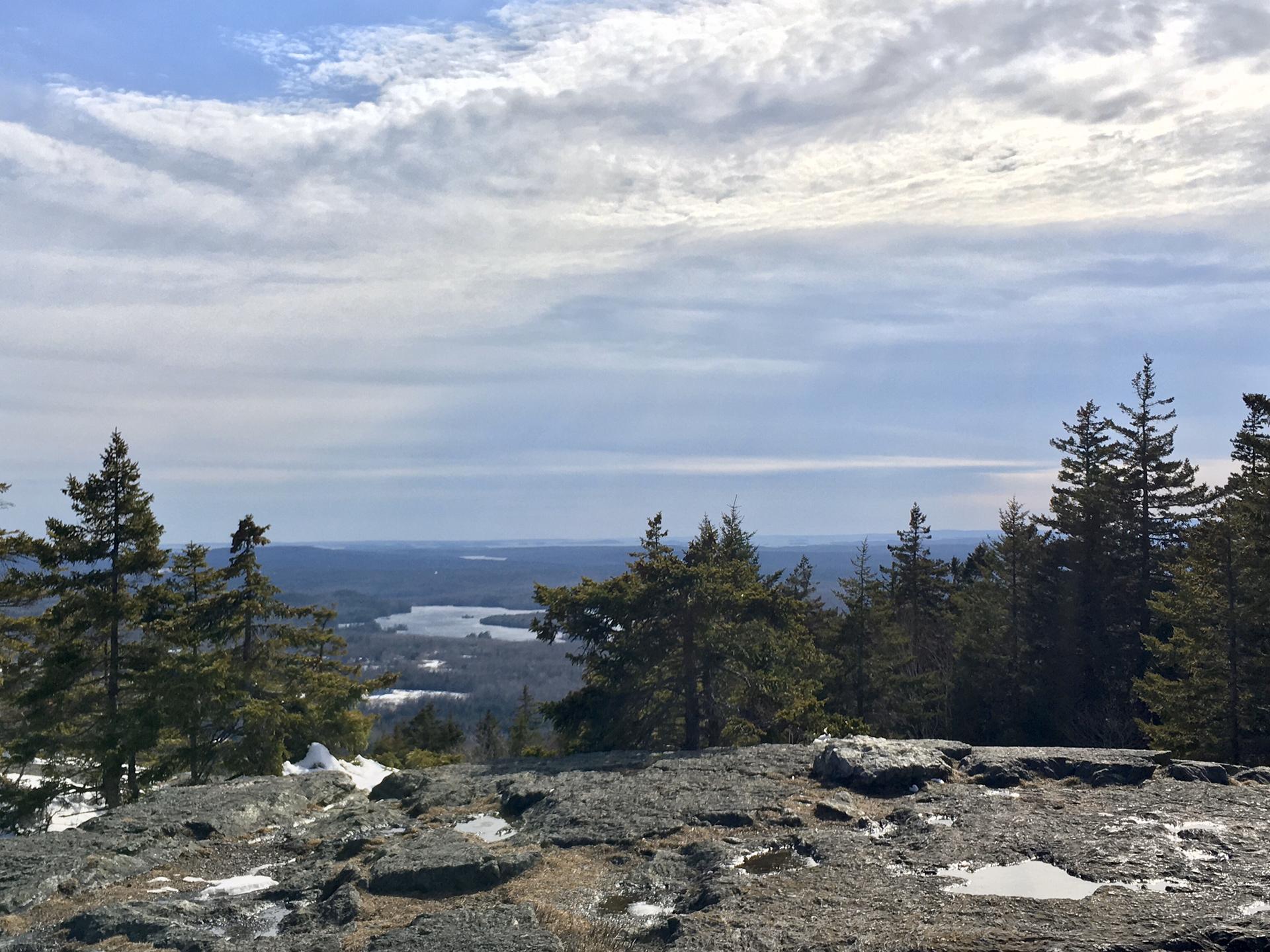 Rocky summit view with evergreen trees, distant lake, and cloudy sky.