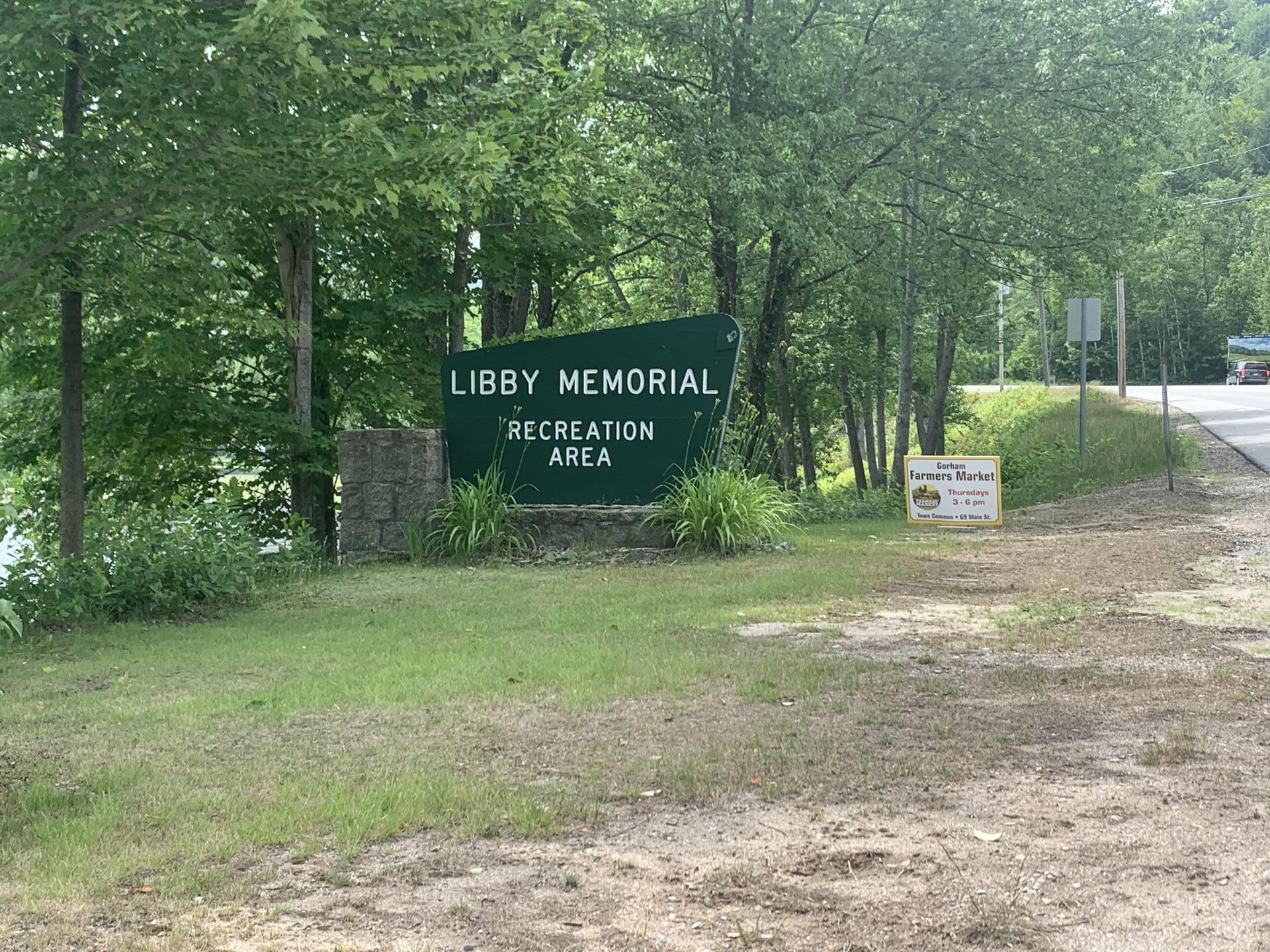 A large green sign in a grassy area surrounded by dense green trees.