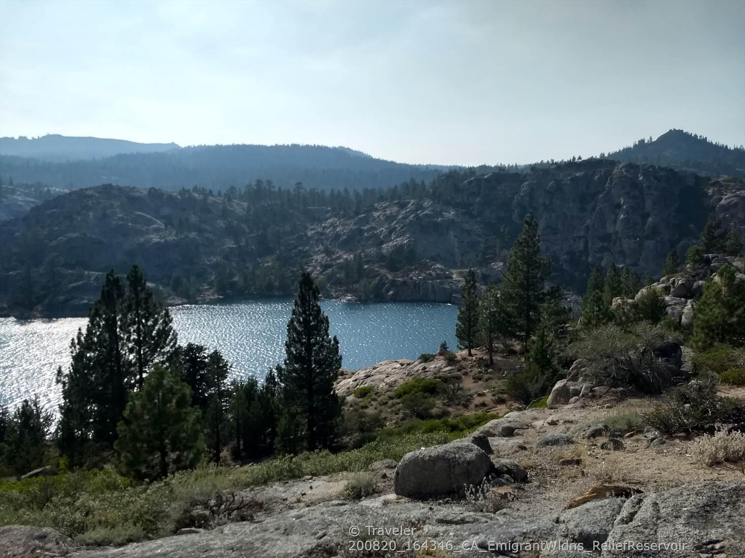 Sparkling lake surrounded by rocky hills and pine trees under a hazy sky.