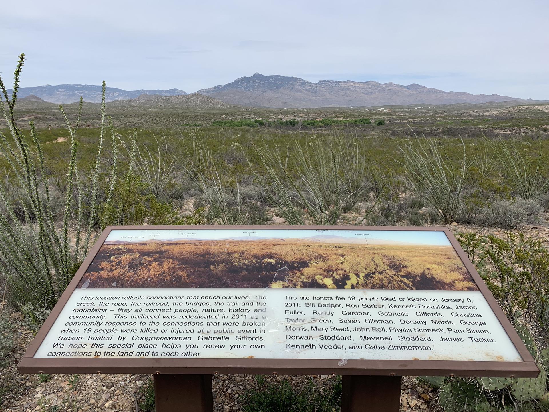 Interpretive sign in desert foreground, with sparse plants and distant mountains under cloudy sky.