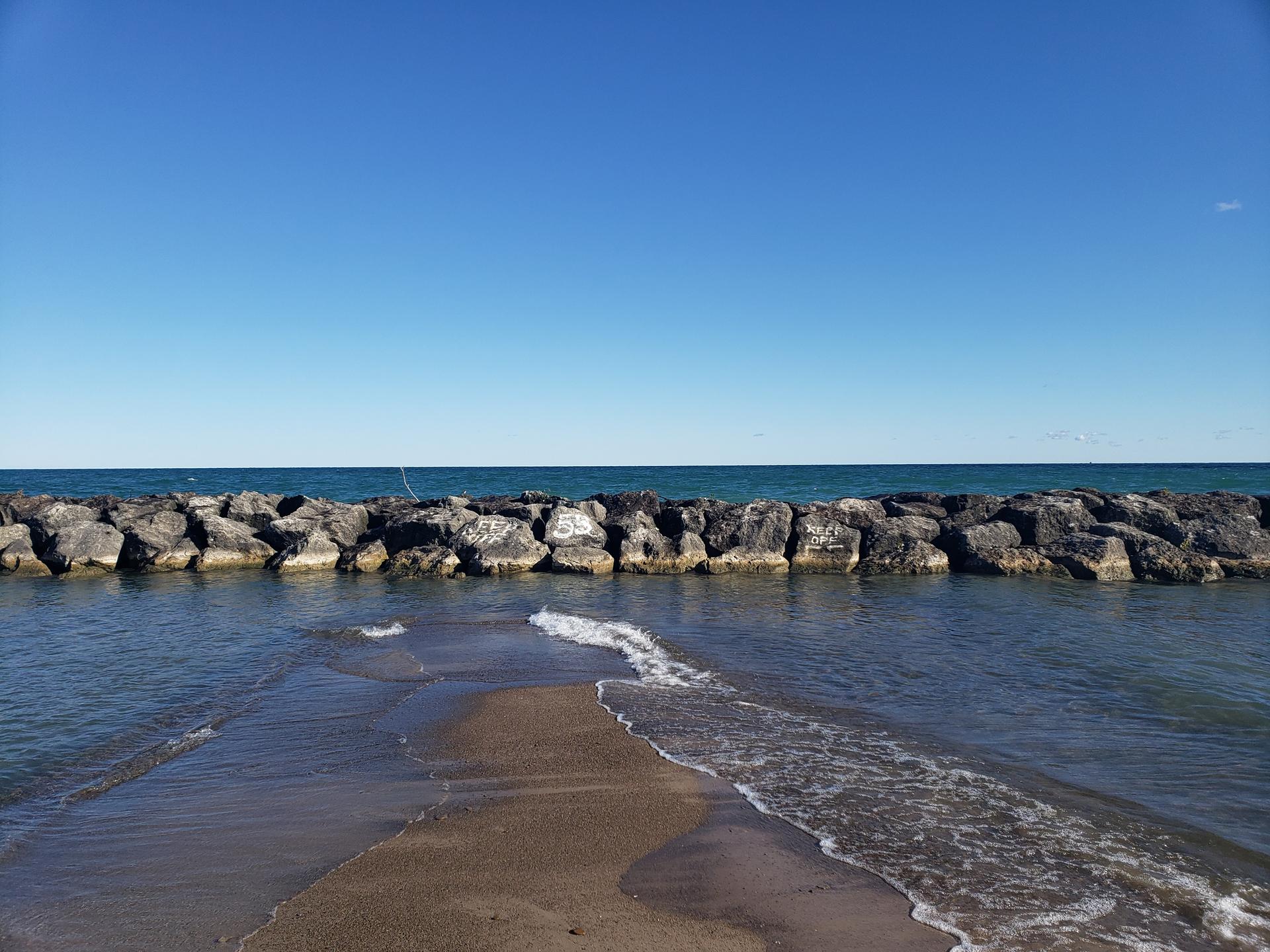 A rocky breakwater extends into calm blue water under a clear sky.