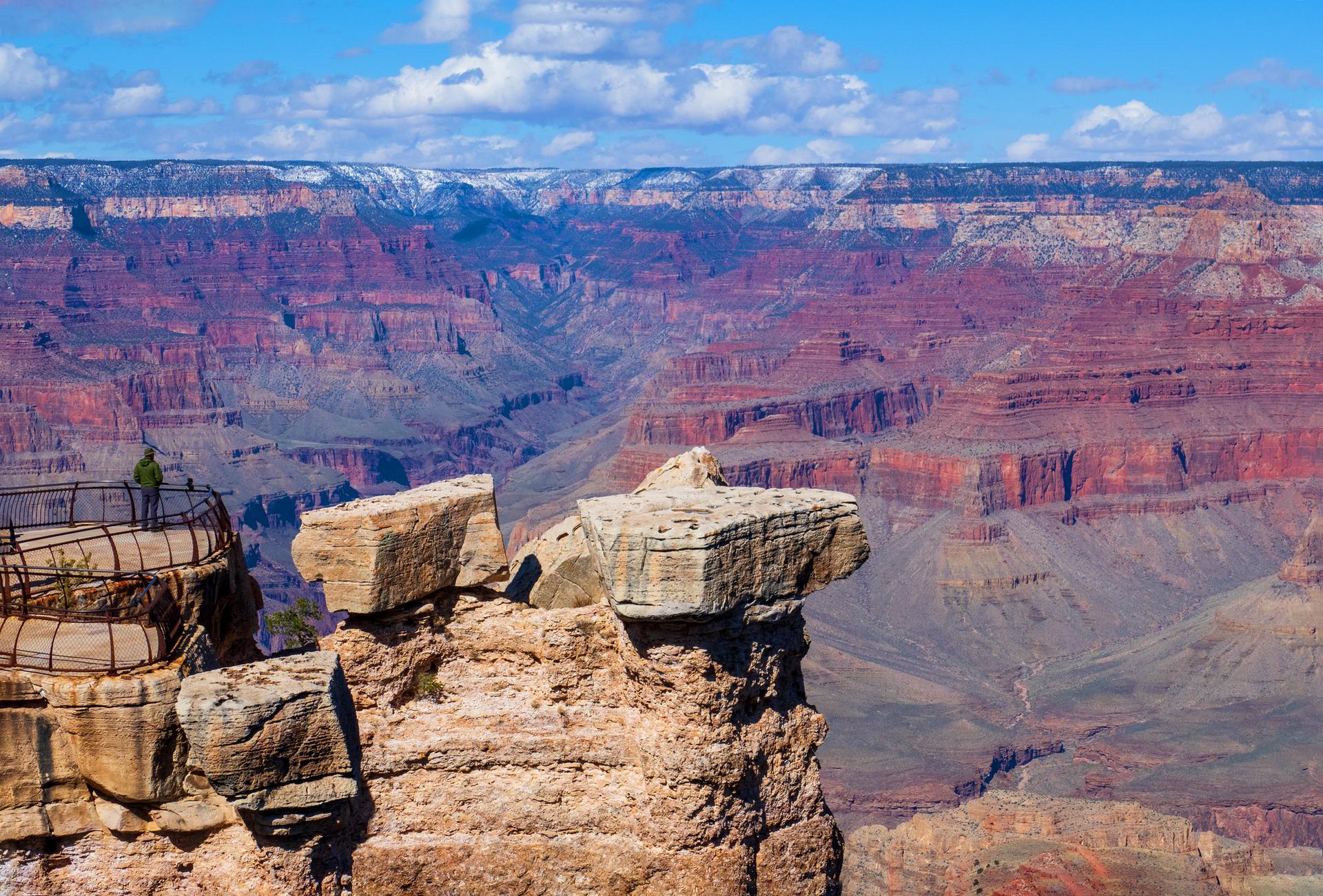 A person on a platform overlooks a vast, colorful canyon under a blue sky.